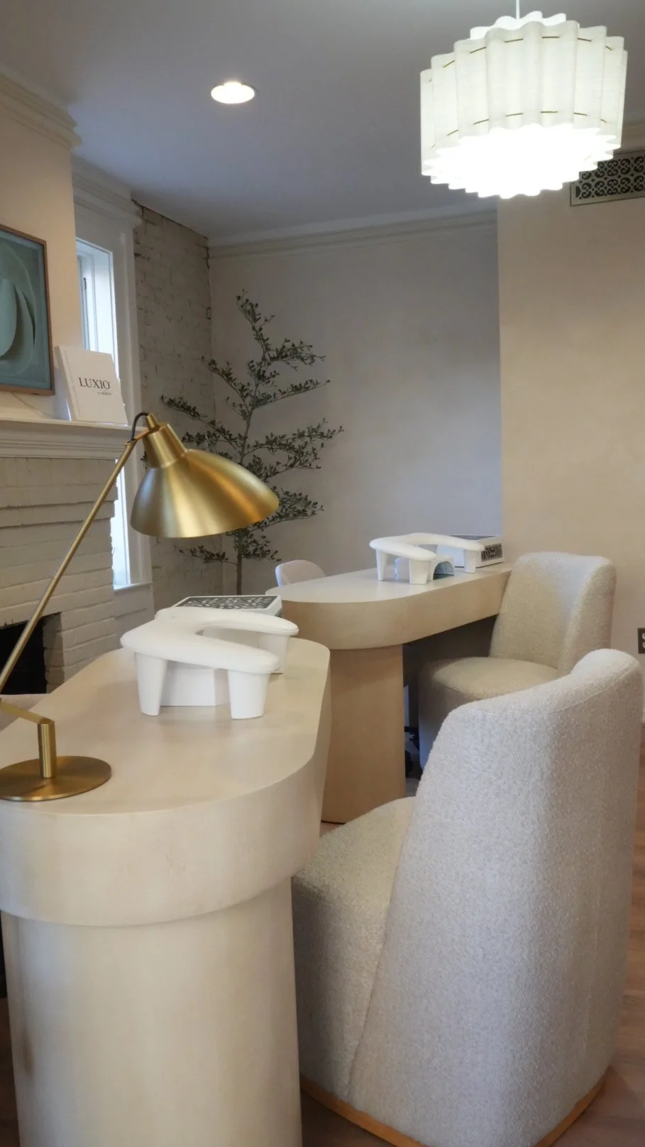Interior of a manicure room with a white brick fireplace, a window, a potted tree, a white desk with a brass desk lamp, and beige upholstered chairs. There are white nail lamps on the desk and a modern white ceiling light fixture.
