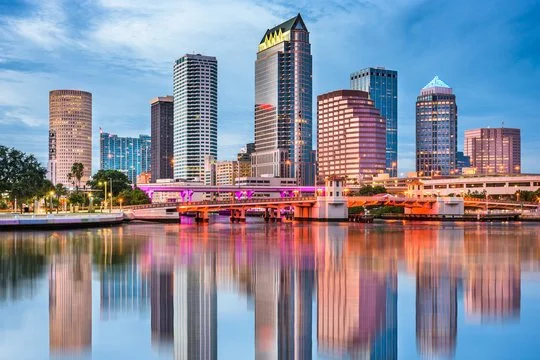 Downtown city skyline with modern high-rise buildings reflection on the water, pink-lit bridge, and a clear sky.