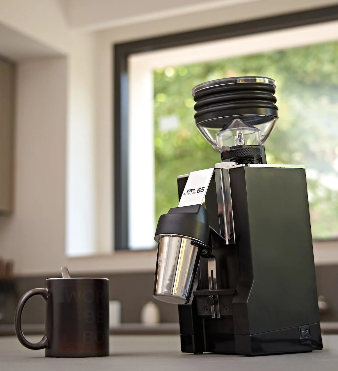 Macchina per il caffè espresso con tazza nera sul tavolo in cucina con finestra a sfondo con alberi verdeggianti.