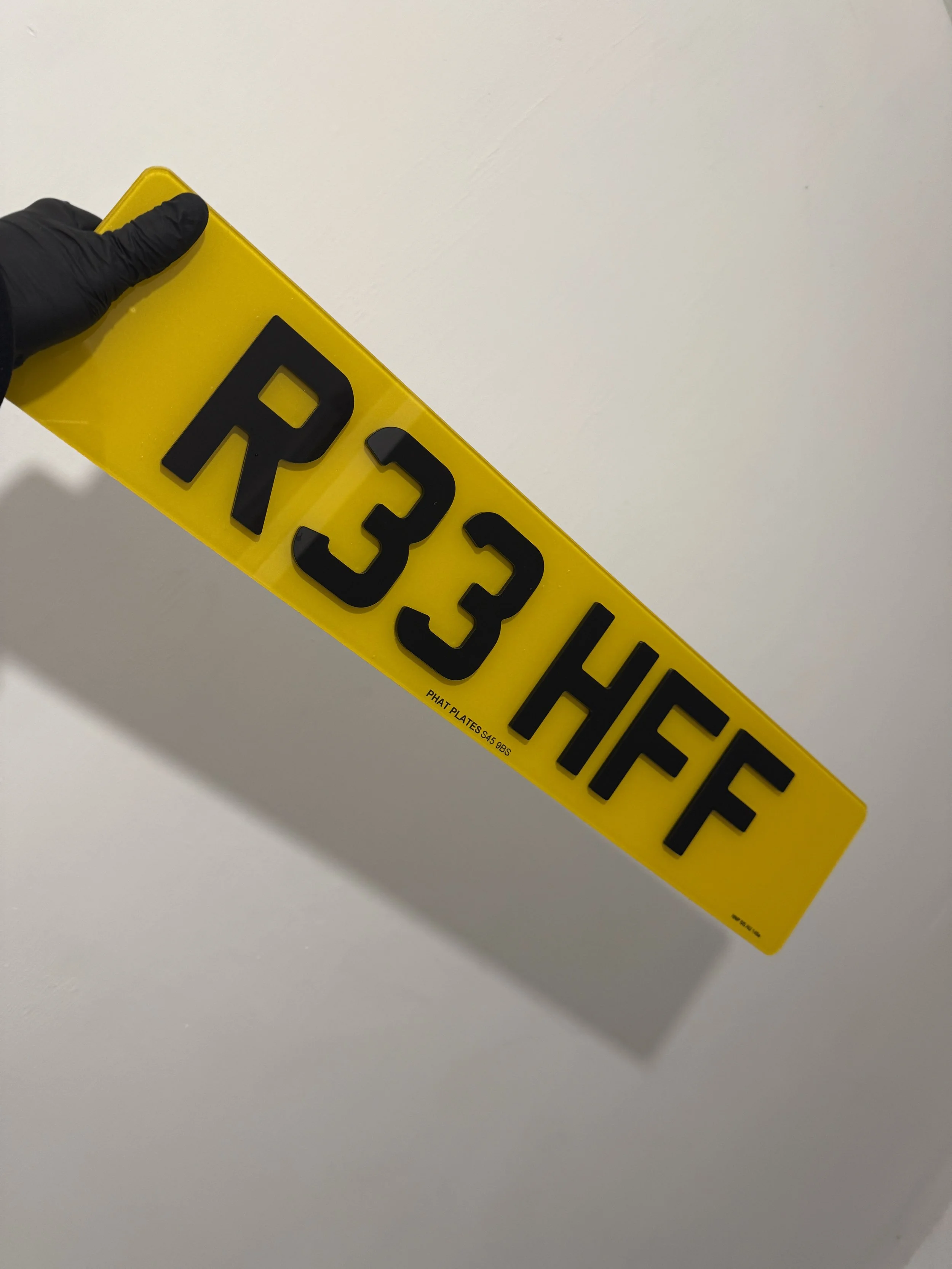 A yellow license plate with black characters reading 'RE33 HFF', held by a hand wearing a black glove, against a plain light-colored background.