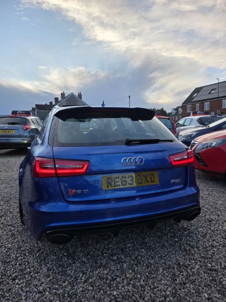 Blue Audi car parked in a gravel lot with other vehicles around it, under a cloudy sky.