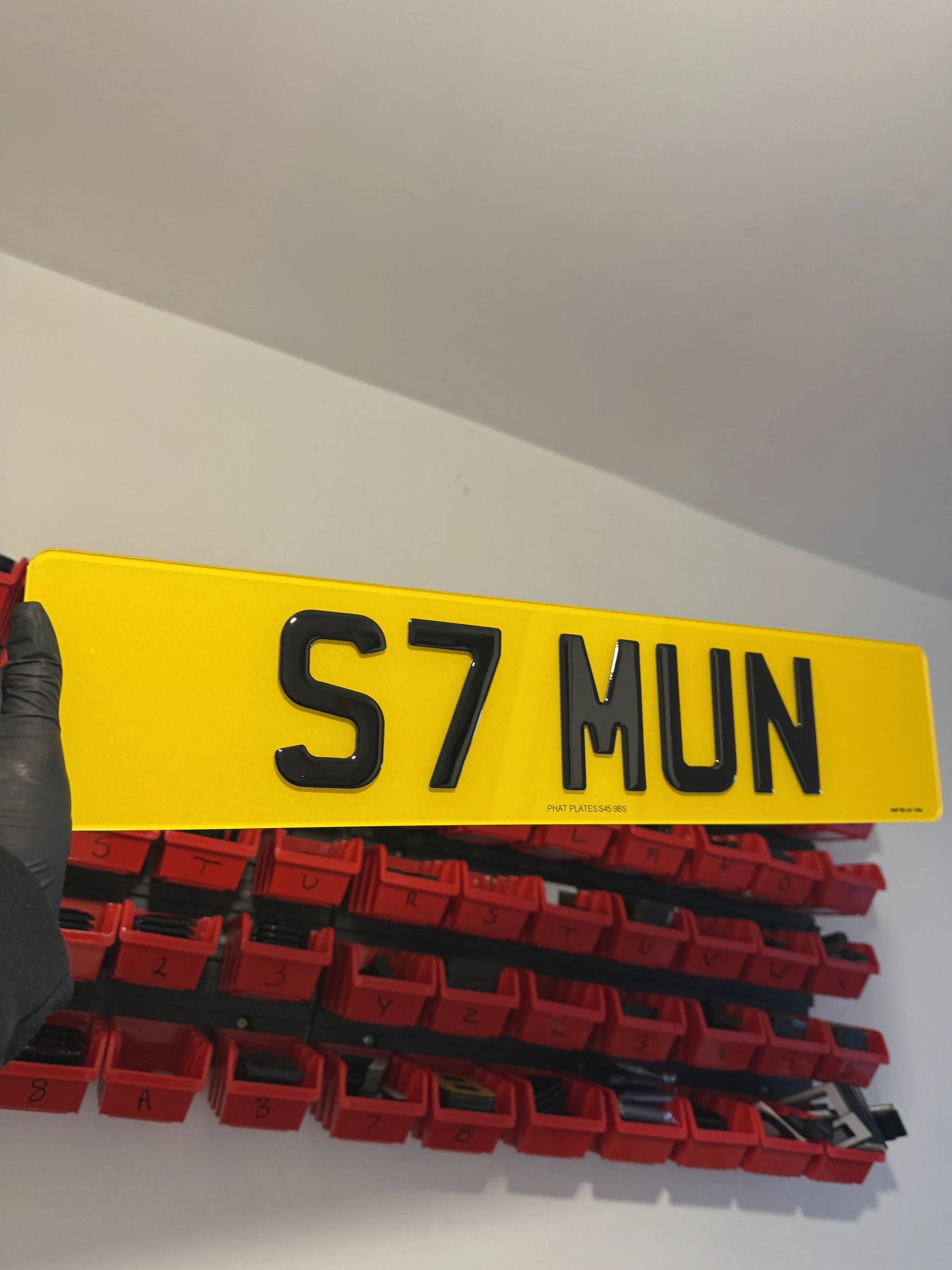 Yellow license plate with black characters reading "S7 MUN" being held by a hand in a black glove, with red tool storage bins containing various small tools or parts in the background.