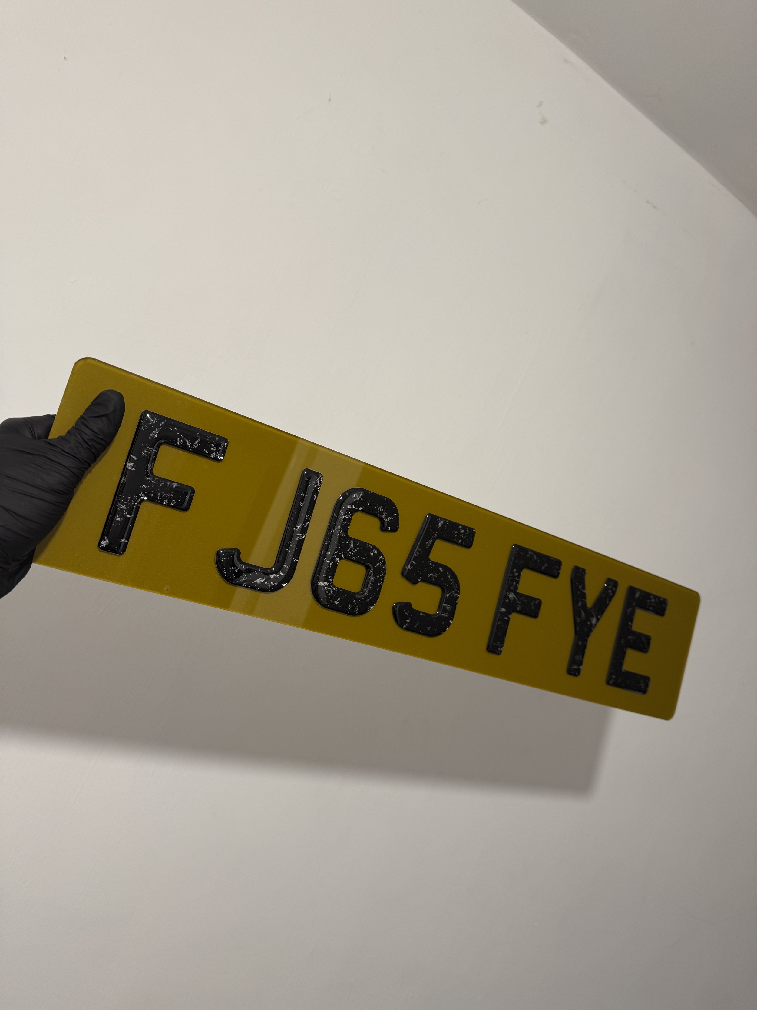 A black-gloved hand holding a yellow reflective license plate with black characters reading 'F J65FYE' against a plain white background.
