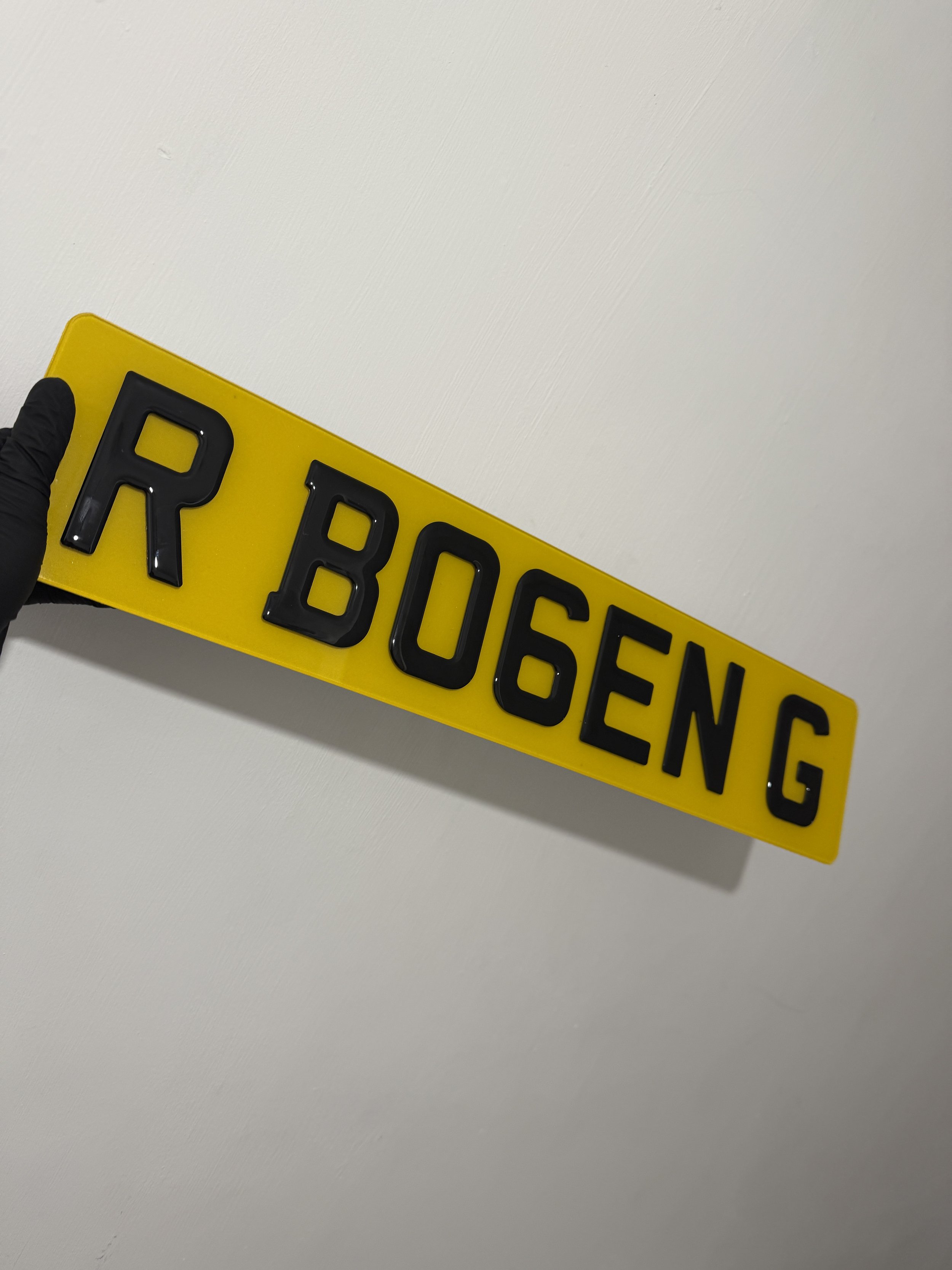 Person wearing a black glove holds a yellow license plate with black letters 'R BOGEN G' against a plain white wall.