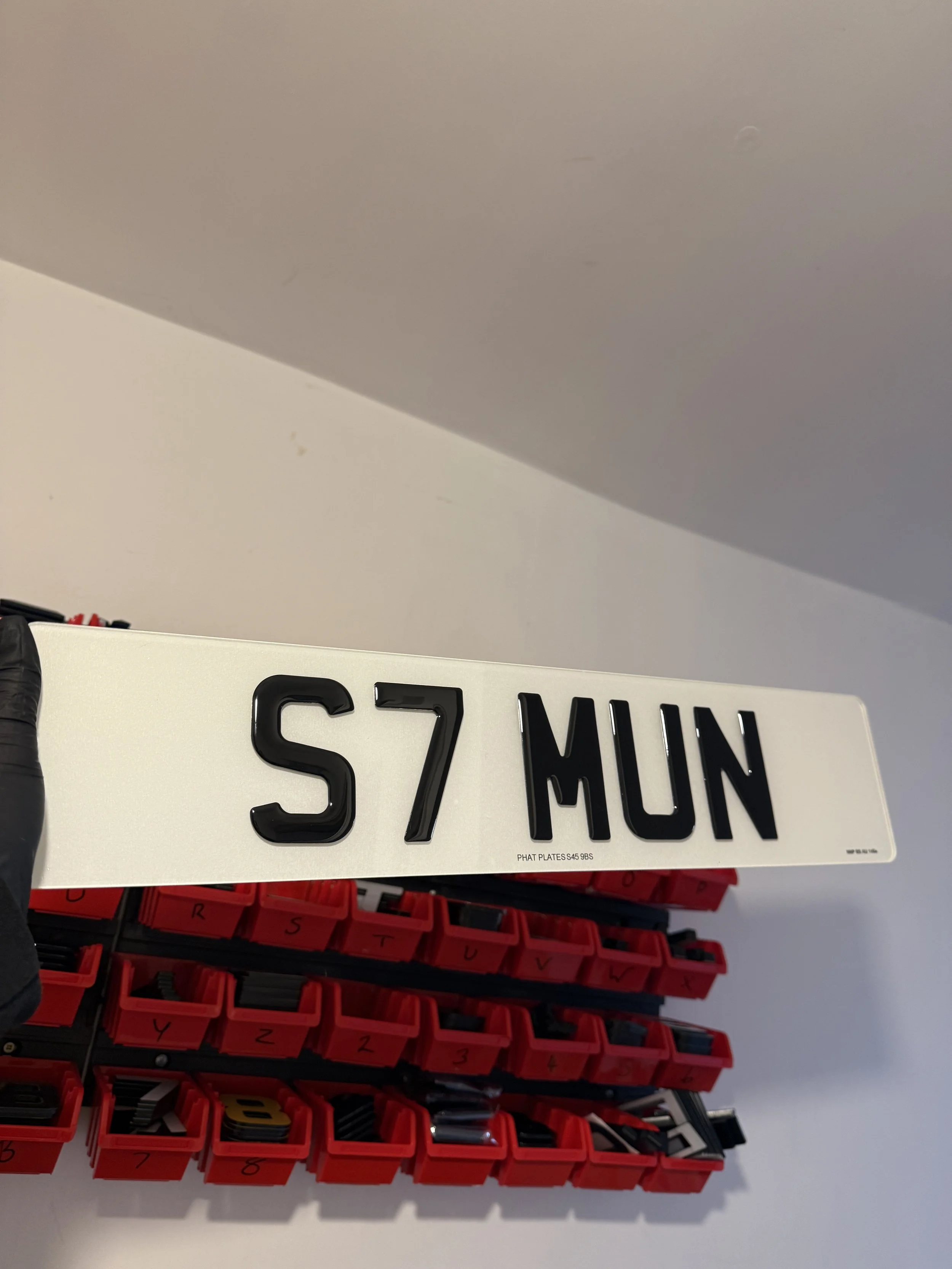 A white license plate with black characters reading "S7 MUN," held by a person wearing a black glove. In the background, there are red and black storage bins with labeled compartments.