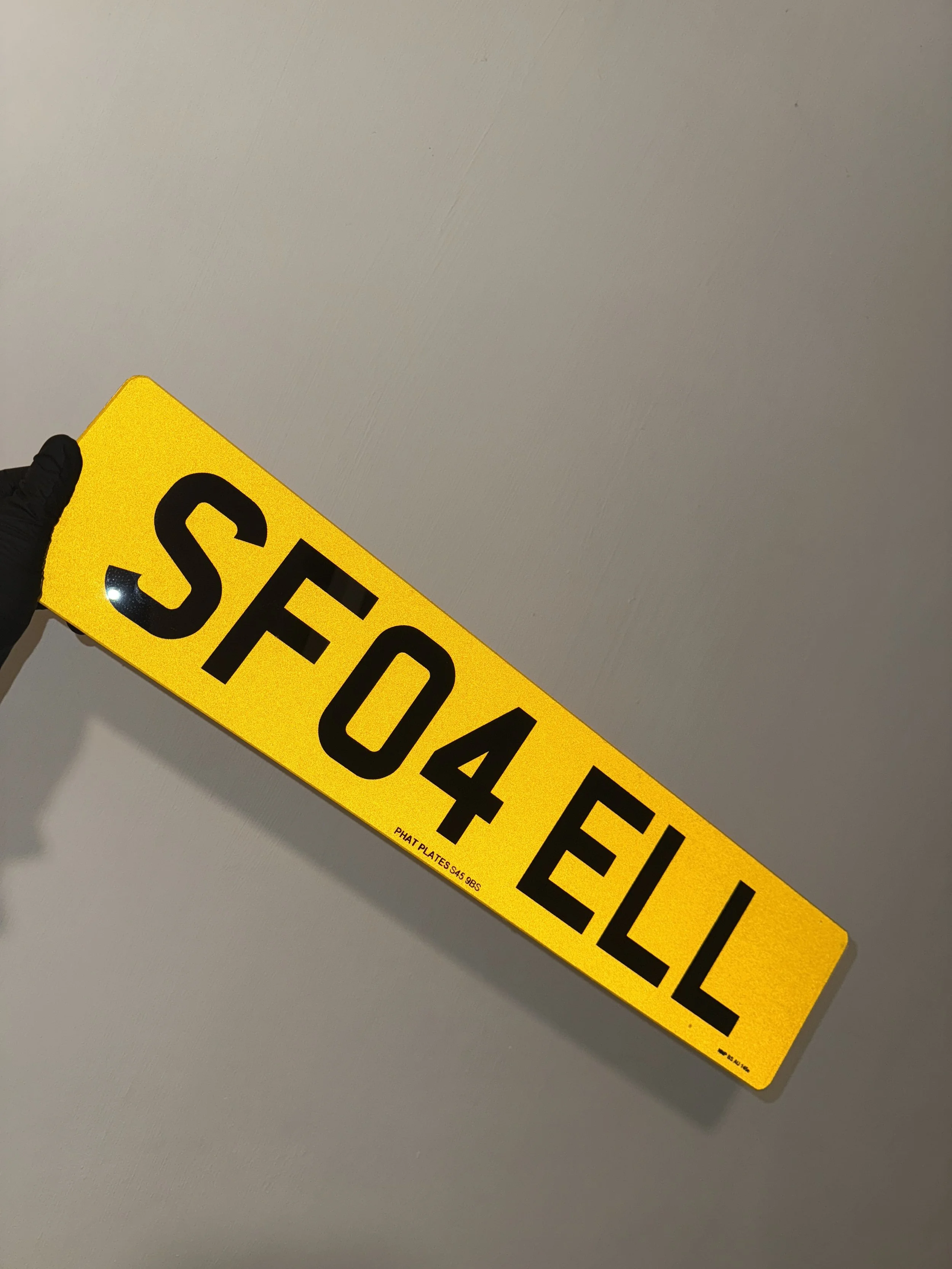 Bright yellow license plate with black letters reading 'SF04 EUL' held by a gloved hand against a plain wall.