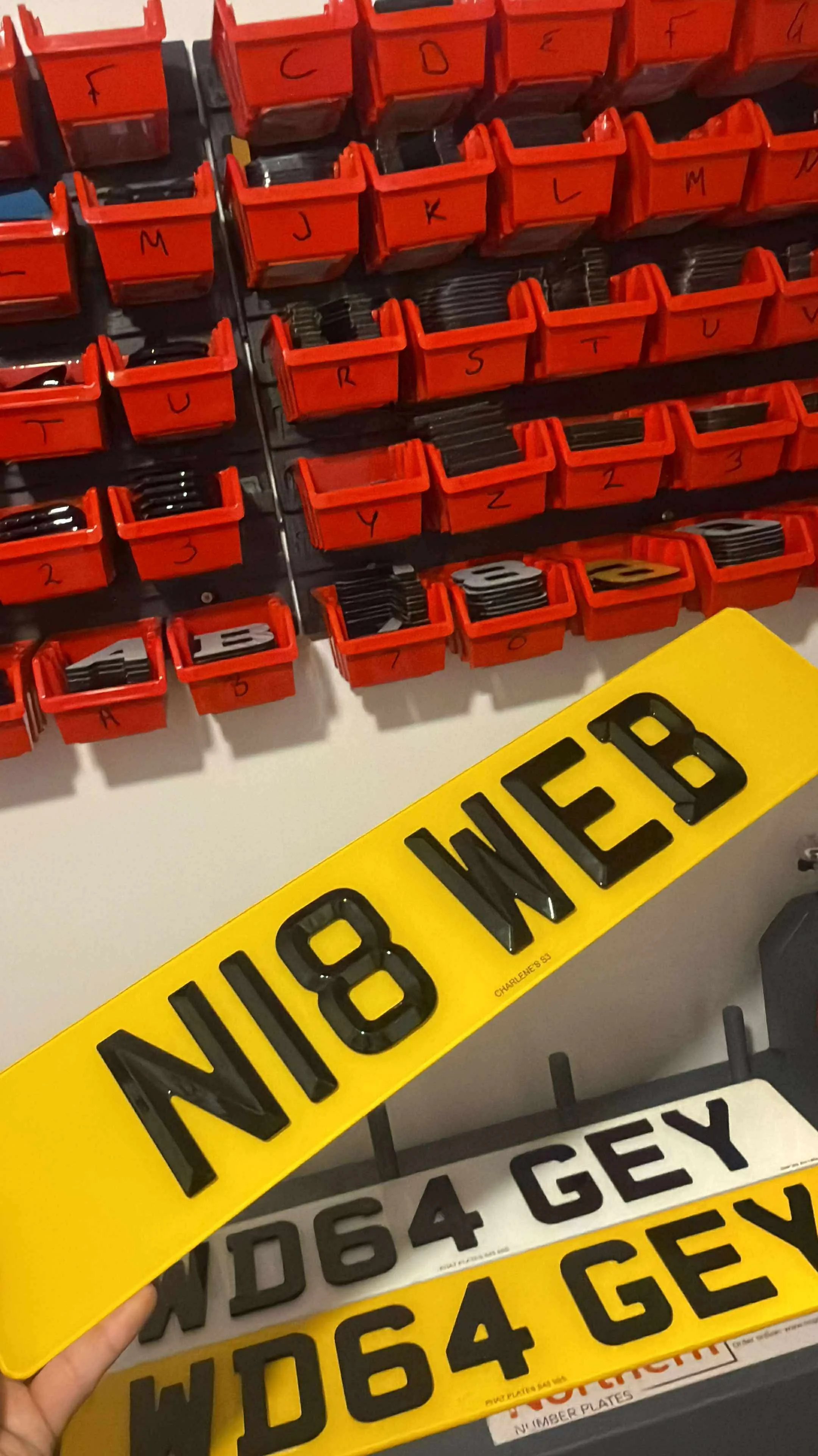 A wall with multiple small red containers labeled with black letters and numbers, and several black and white license plates, including a yellow license plate that reads 'N18 WEB'.