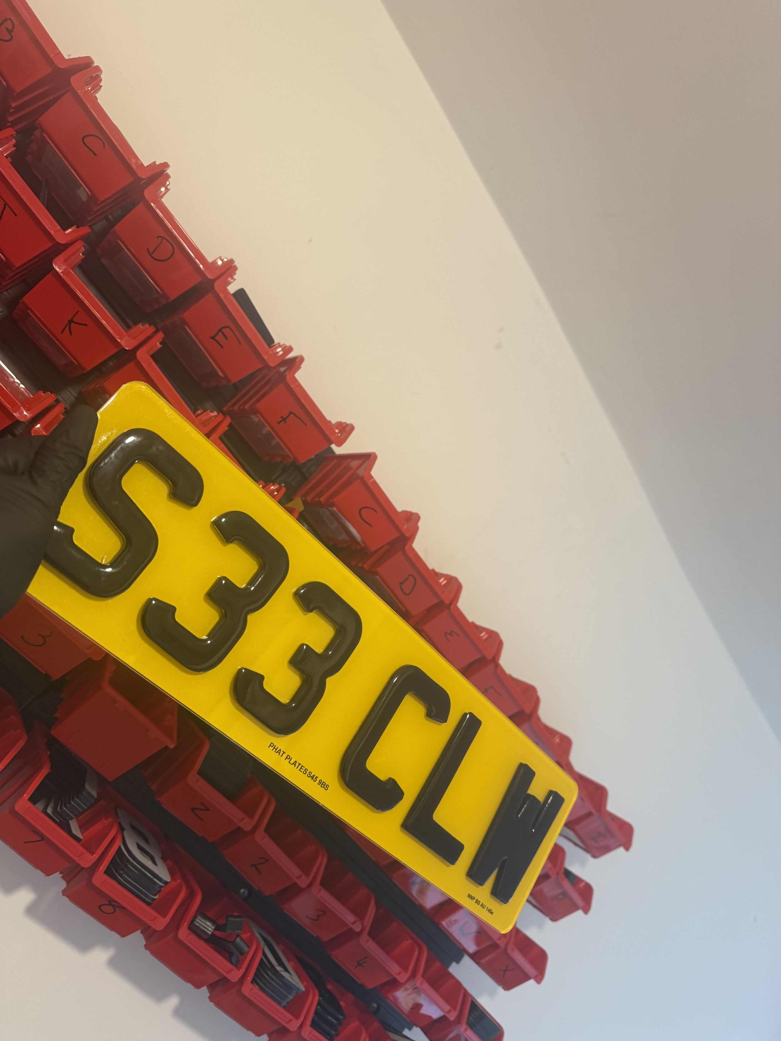Close-up of a yellow license plate with black numbers and letters, held by a hand wearing a black glove, against background of red labeled storage bins.