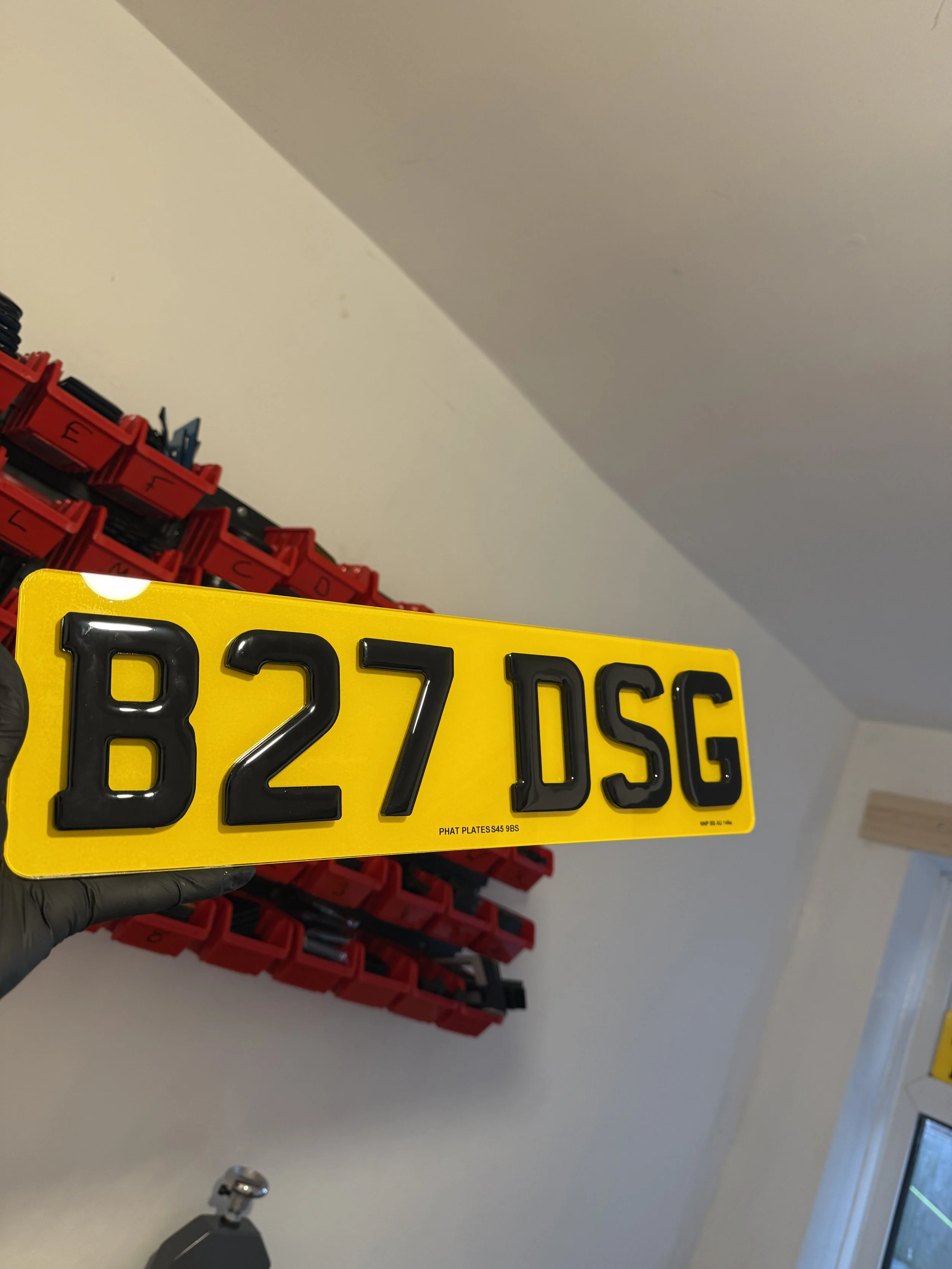 A yellow European-style license plate with black characters reading 'B27 DSG' held by a person wearing a black glove. There are red plastic containers stacked in the background against a white wall and a slanted ceiling.
