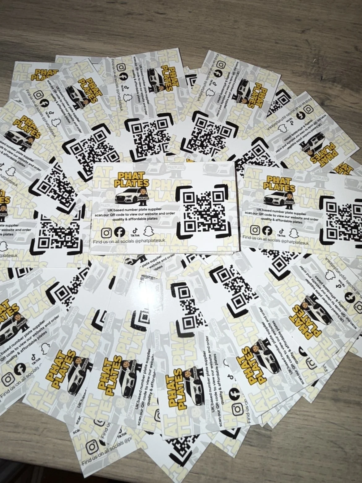A pile of business cards for Phat Plates, a UK-based number plate supplier, featuring QR codes, social media icons, and a cartoon character.