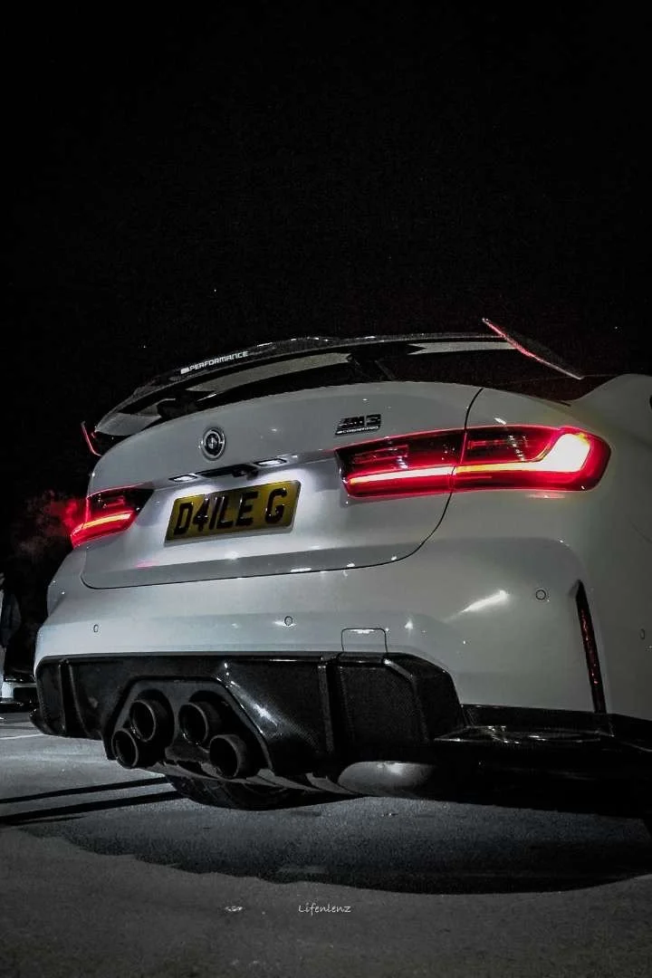 Rear view of a white BMW M3 car at night, showing quad exhaust pipes, distinctive taillights, and a custom license plate 'D4LE C'.