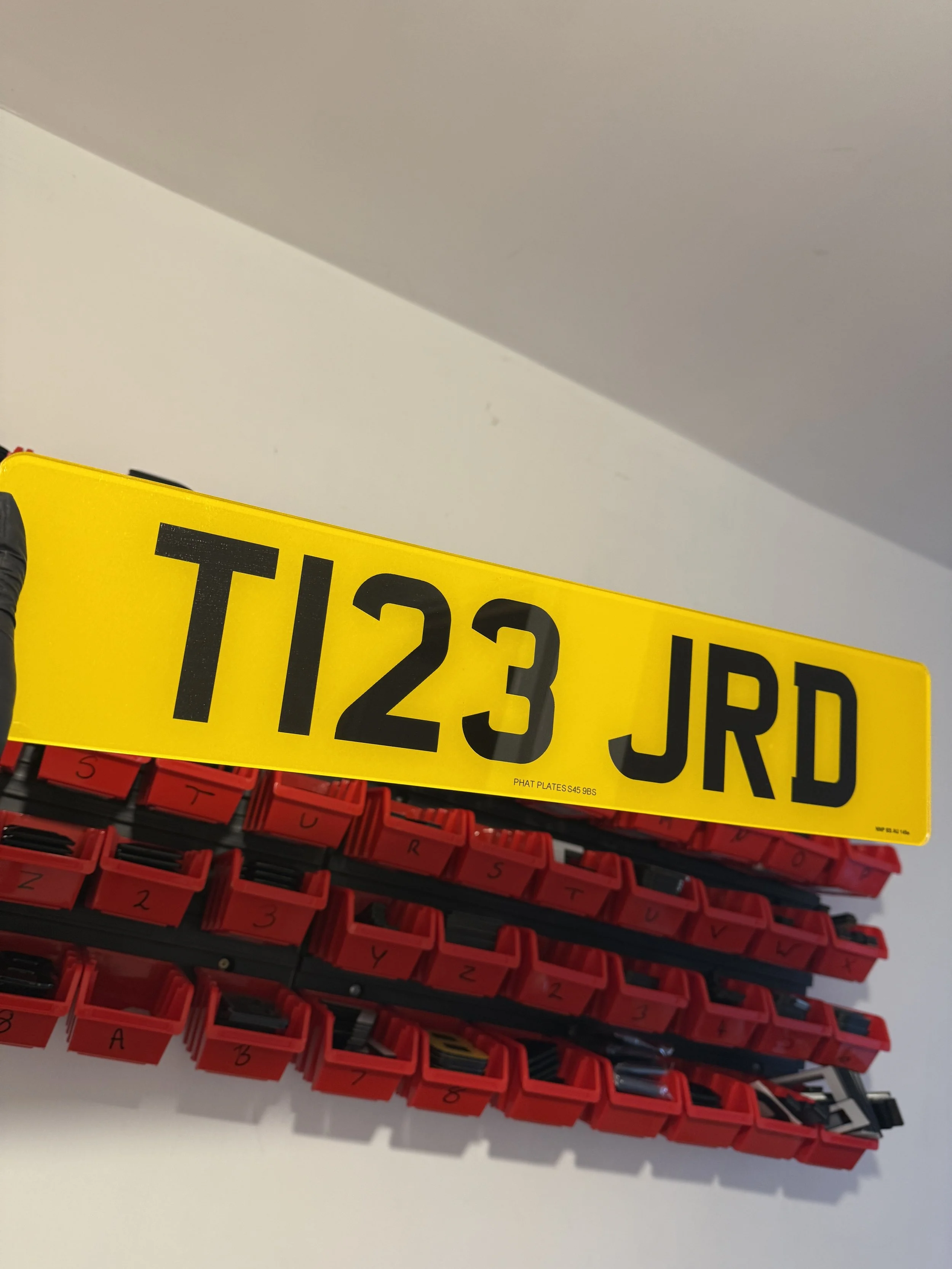 A yellow road sign with the text 'T123 JRD' in black on top of a collection of red containers with small labeled compartments.
