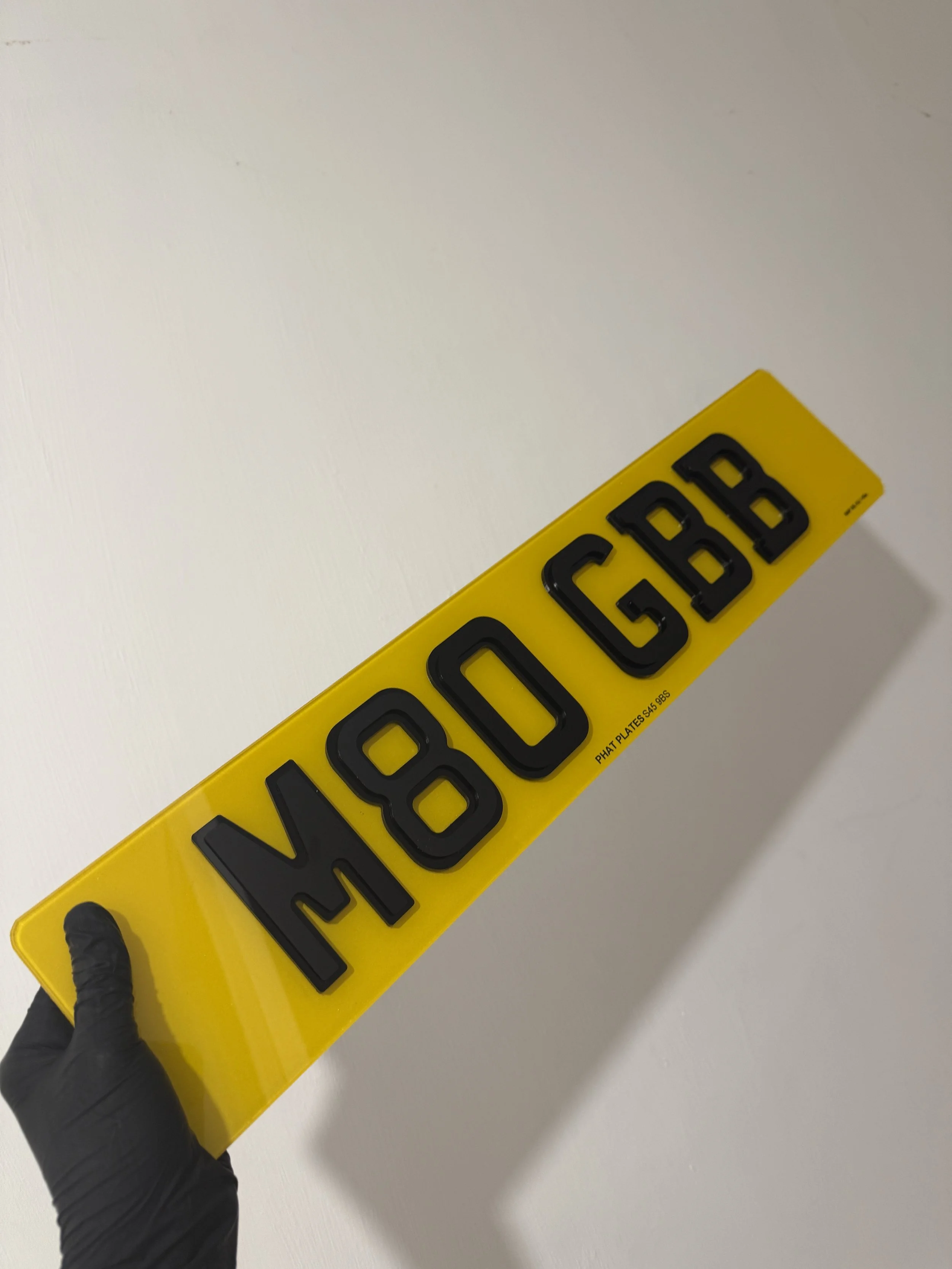A yellow license plate with black raised letters reading "H80 CBD," being held by a hand wearing a black glove, against a plain white wall.