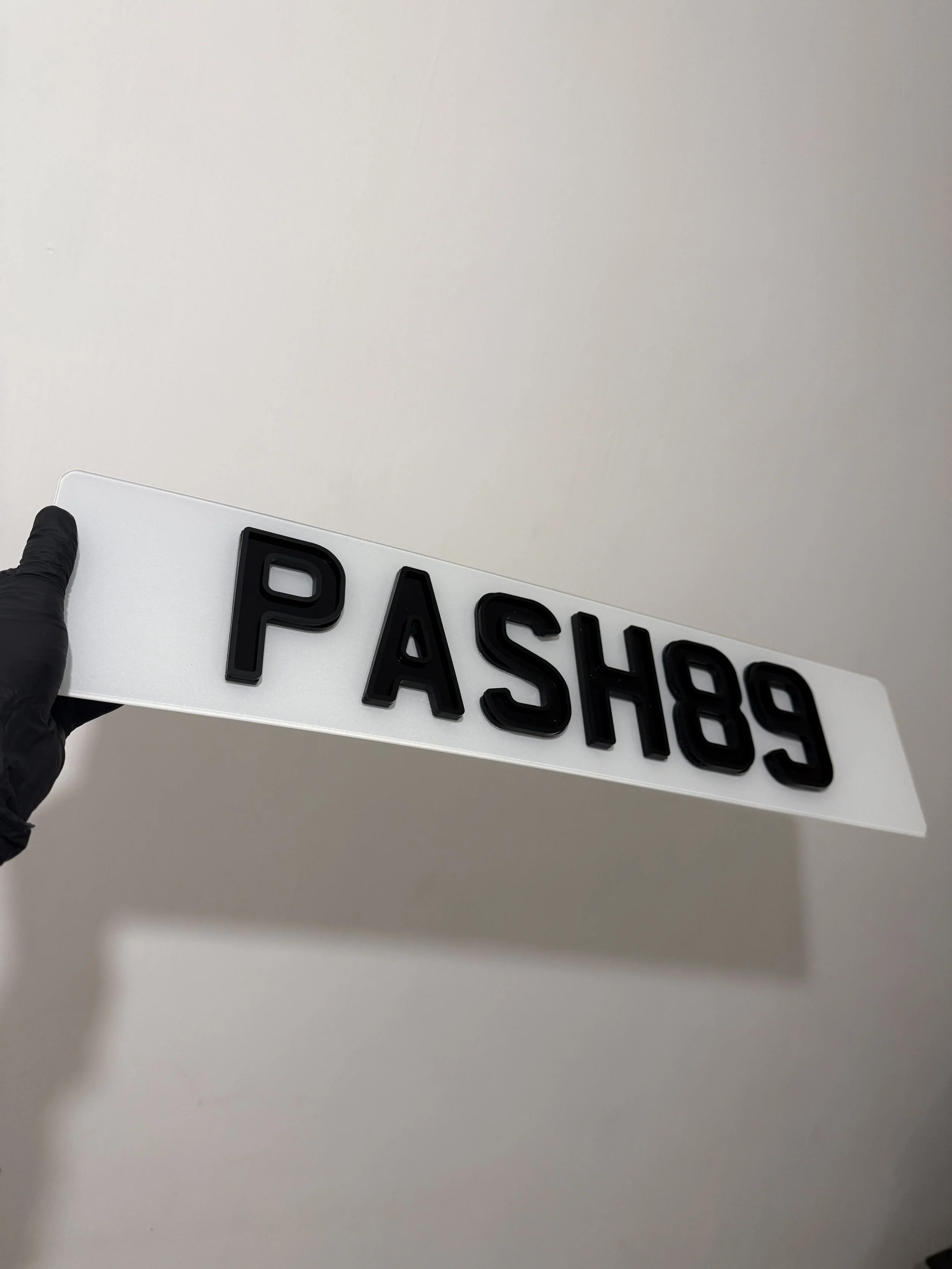 Hand holding a black and white sign with the letters and numbers 'PASHE9' against a plain wall.