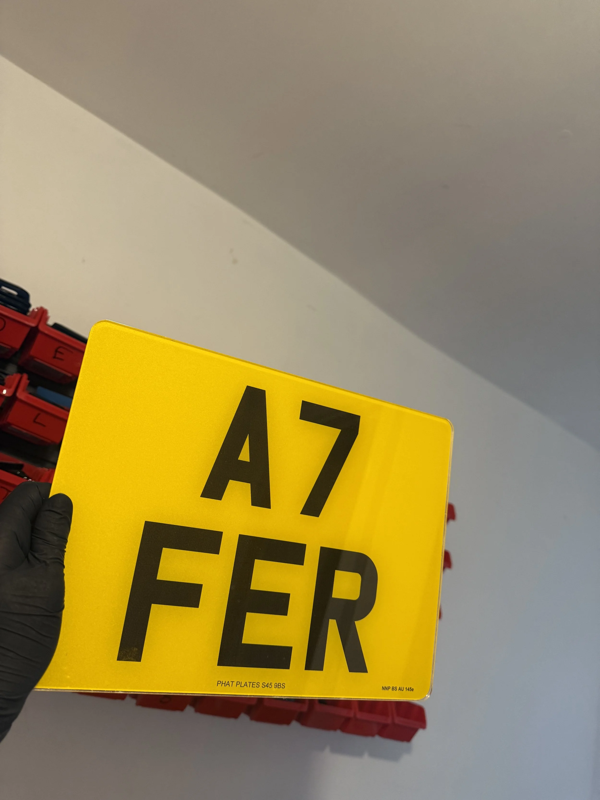 A person wearing a black glove holding a yellow license plate with the characters 'A7 FÉR' against a background of stacked red containers or bins.