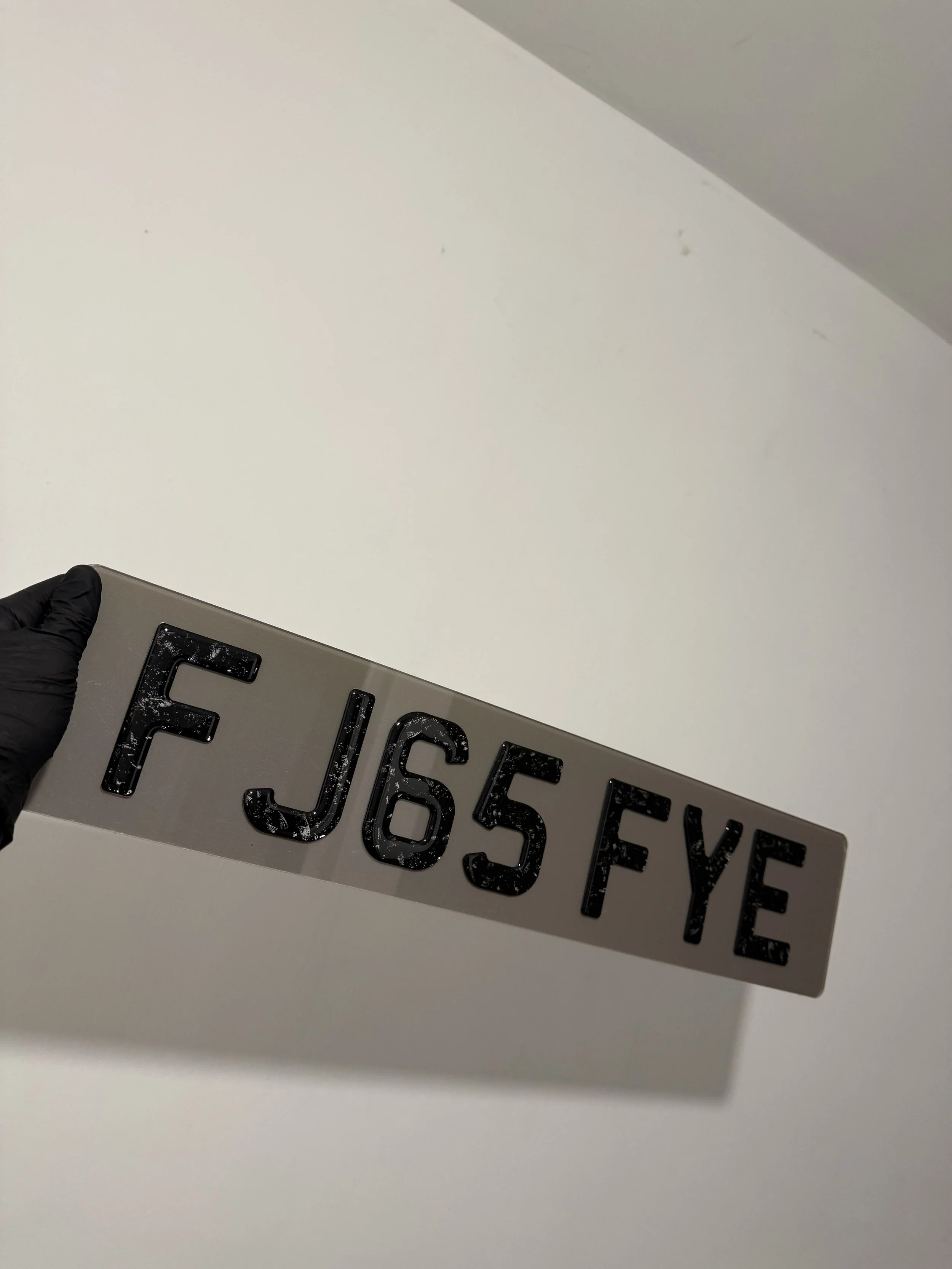 A close-up of a rectangular sign with black letters and numbers reading 'F J65 FYE' being held by a person wearing a black glove, against a white wall background.
