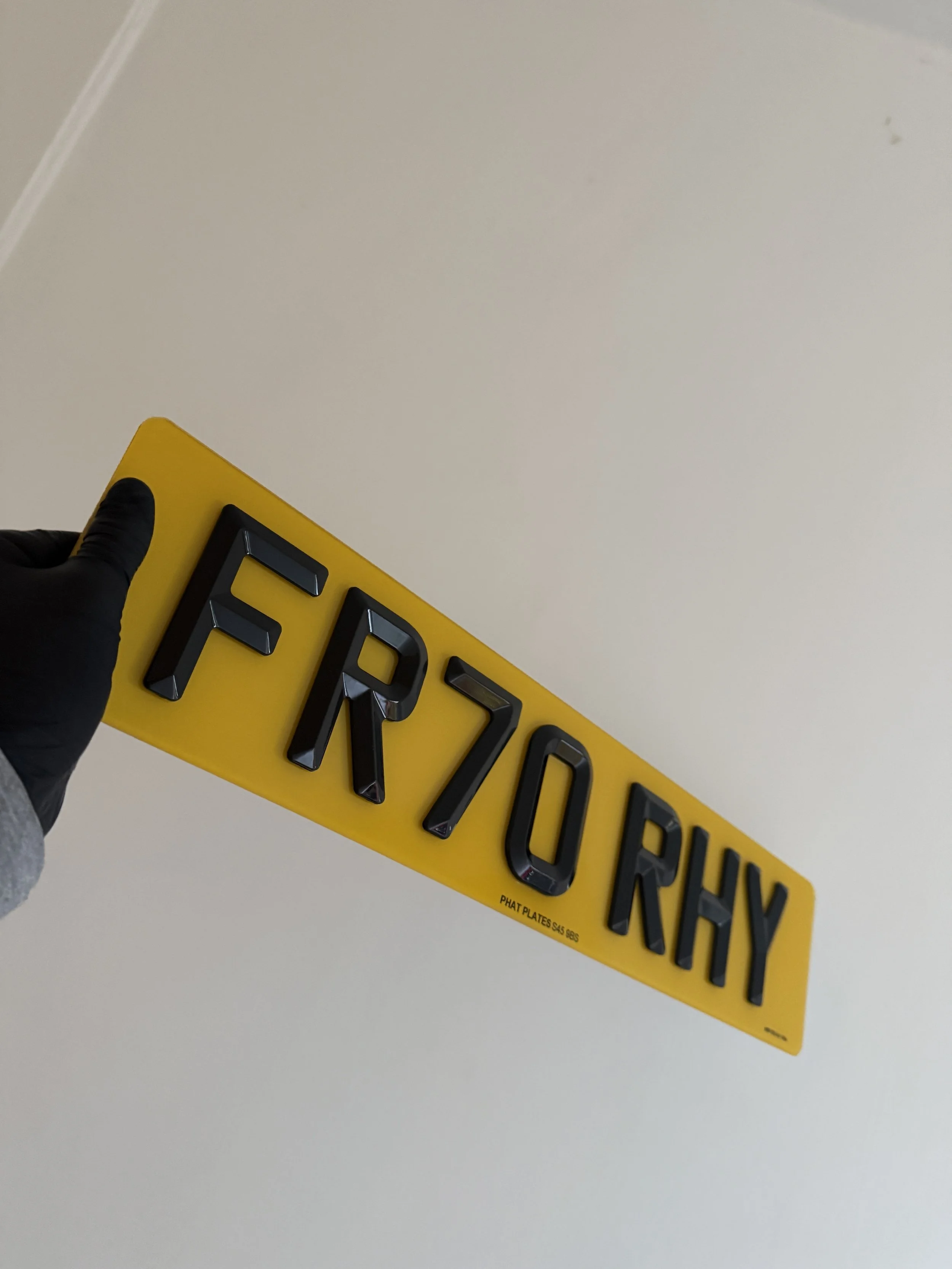 A black-gloved hand holding a yellow license plate with the characters 'FRZ0 RHY' in black 3D letters against a plain white wall background.