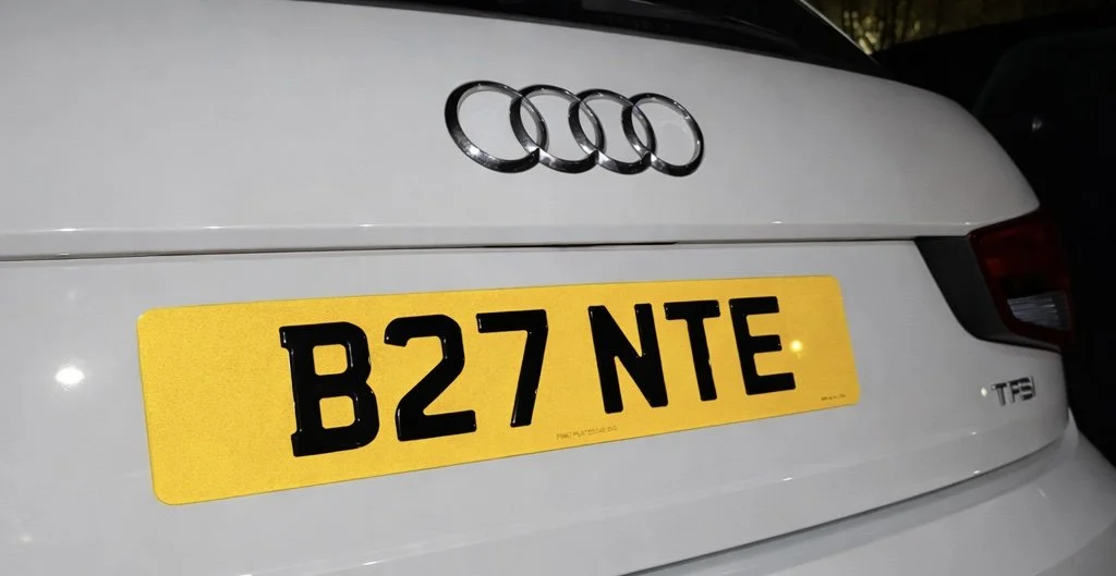 The rear of a white Audi car showing the four-ring logo, a yellow UK license plate with the registration B27 NTE, and part of the taillight.