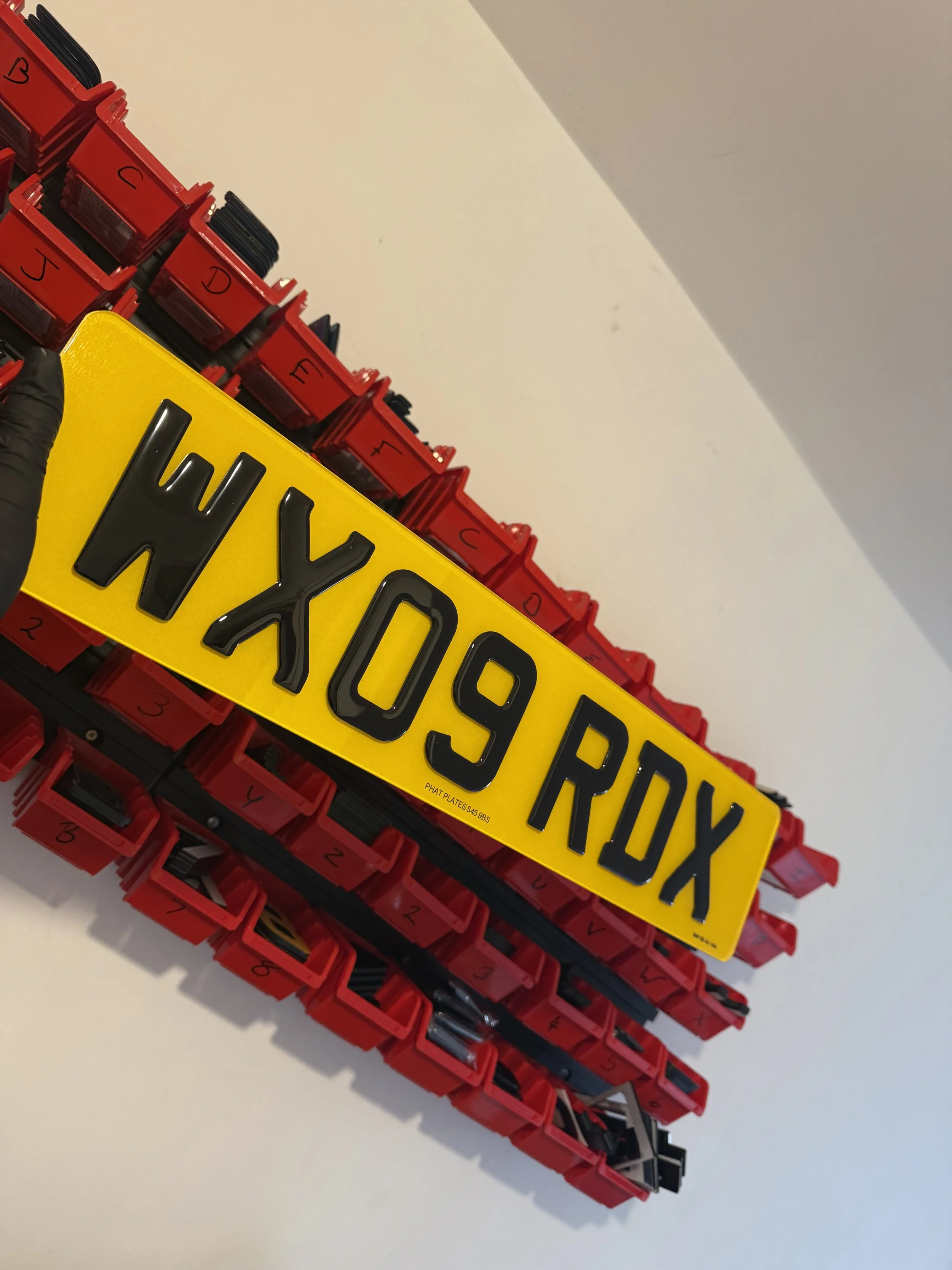 A yellow license plate with black characters resting on top of red plastic organizer drawers filled with various items. The license plate reads 'HX09ROX'.