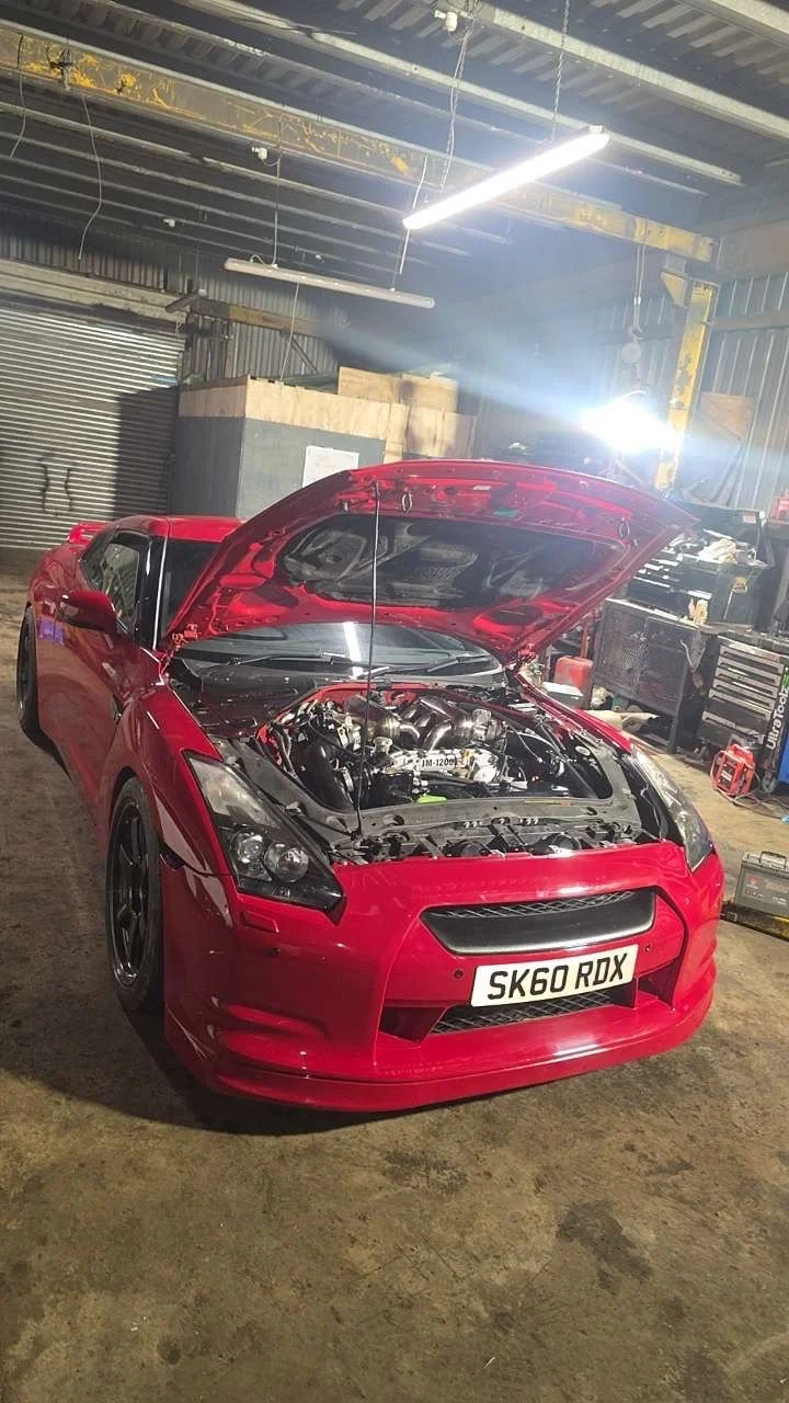 Red sports car with its hood open inside a garage or workshop with tools and equipment around.