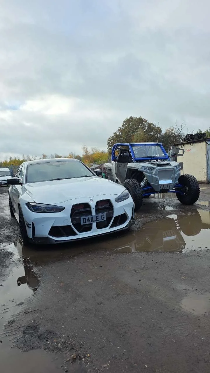 A white modern sports car and a partially assembled or modified off-road vehicle are parked on a muddy surface with puddles, under a cloudy sky.