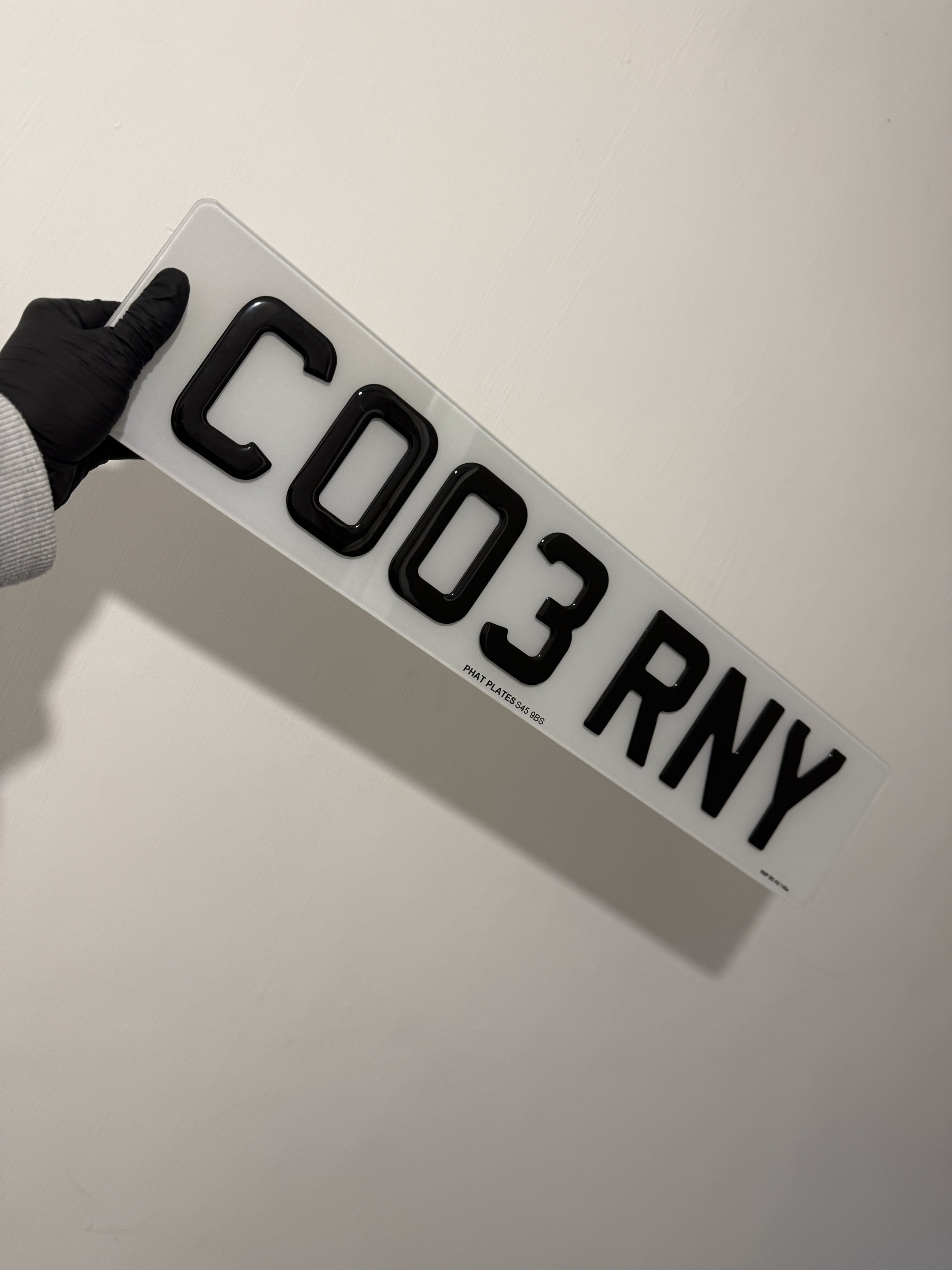 A person wearing a black glove and a gray sweater holding a sign with black illuminated characters that read 'COO3 RNY' against a plain white wall background.