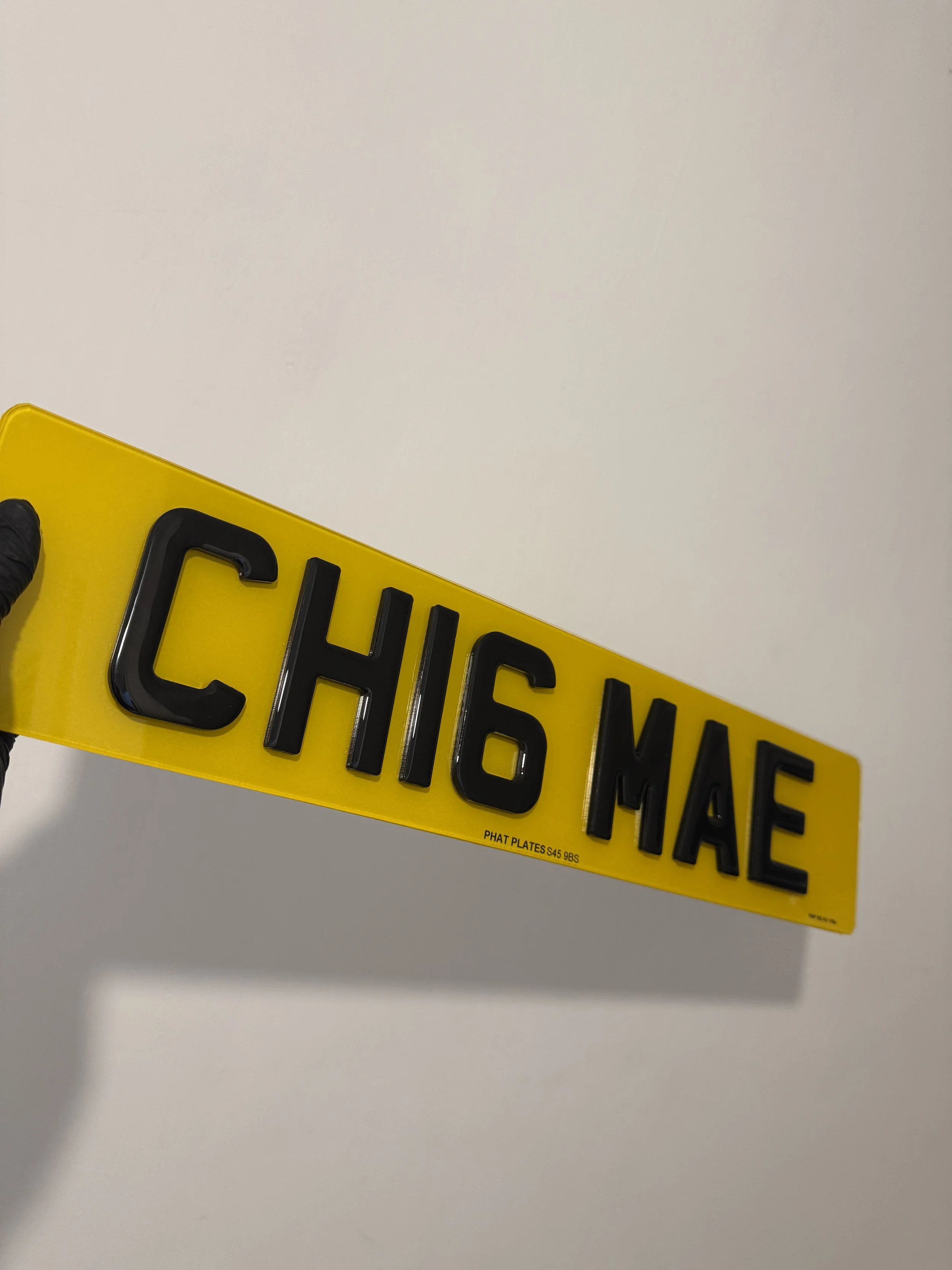 A yellow license plate with black 3D letters reading 'CH16 MAE' being held by a person wearing a black glove against a plain white background.