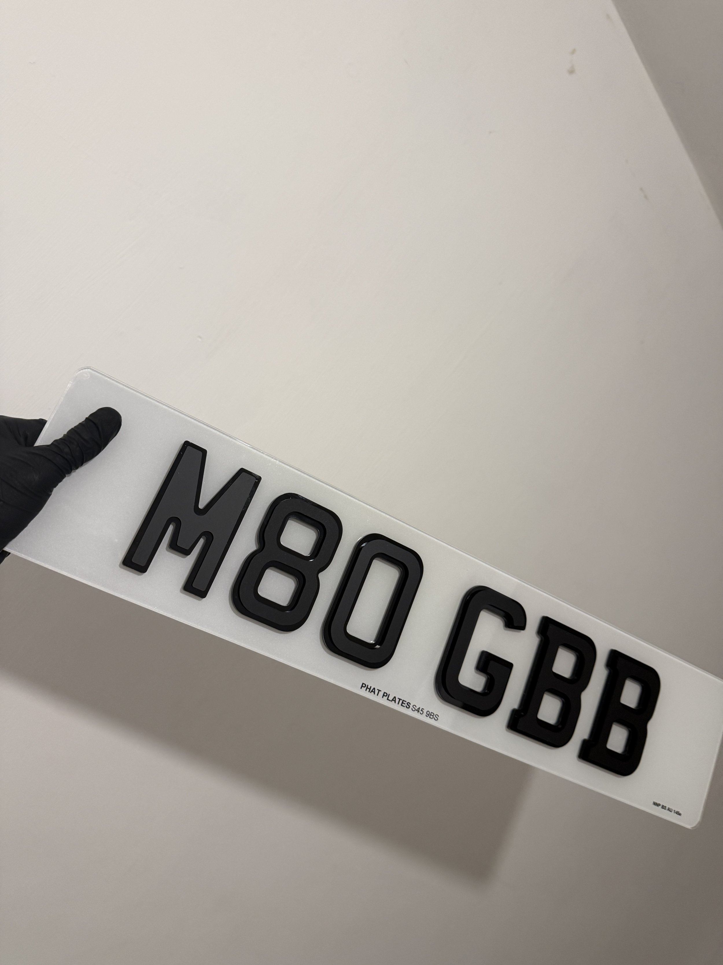 Sign with black raised letters on a white background spelling 'M80 GBB' being held by a hand wearing a black glove.