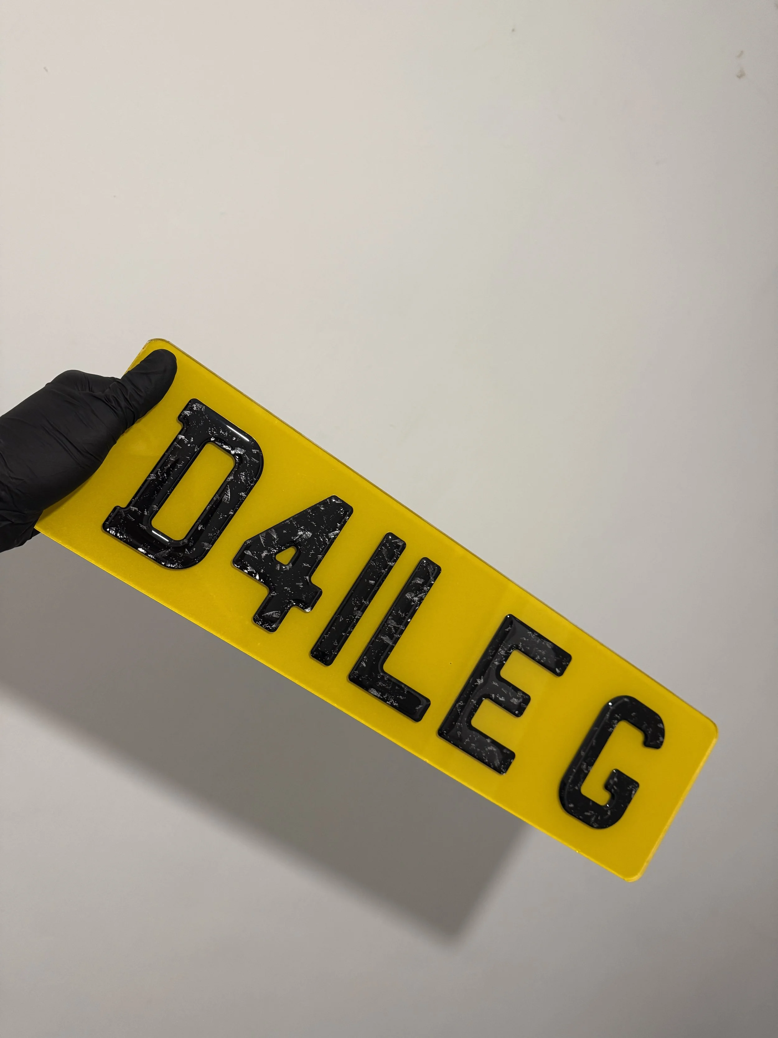 Yellow license plate with black embossed characters 'D4LÉG' being held by a hand wearing a black glove.