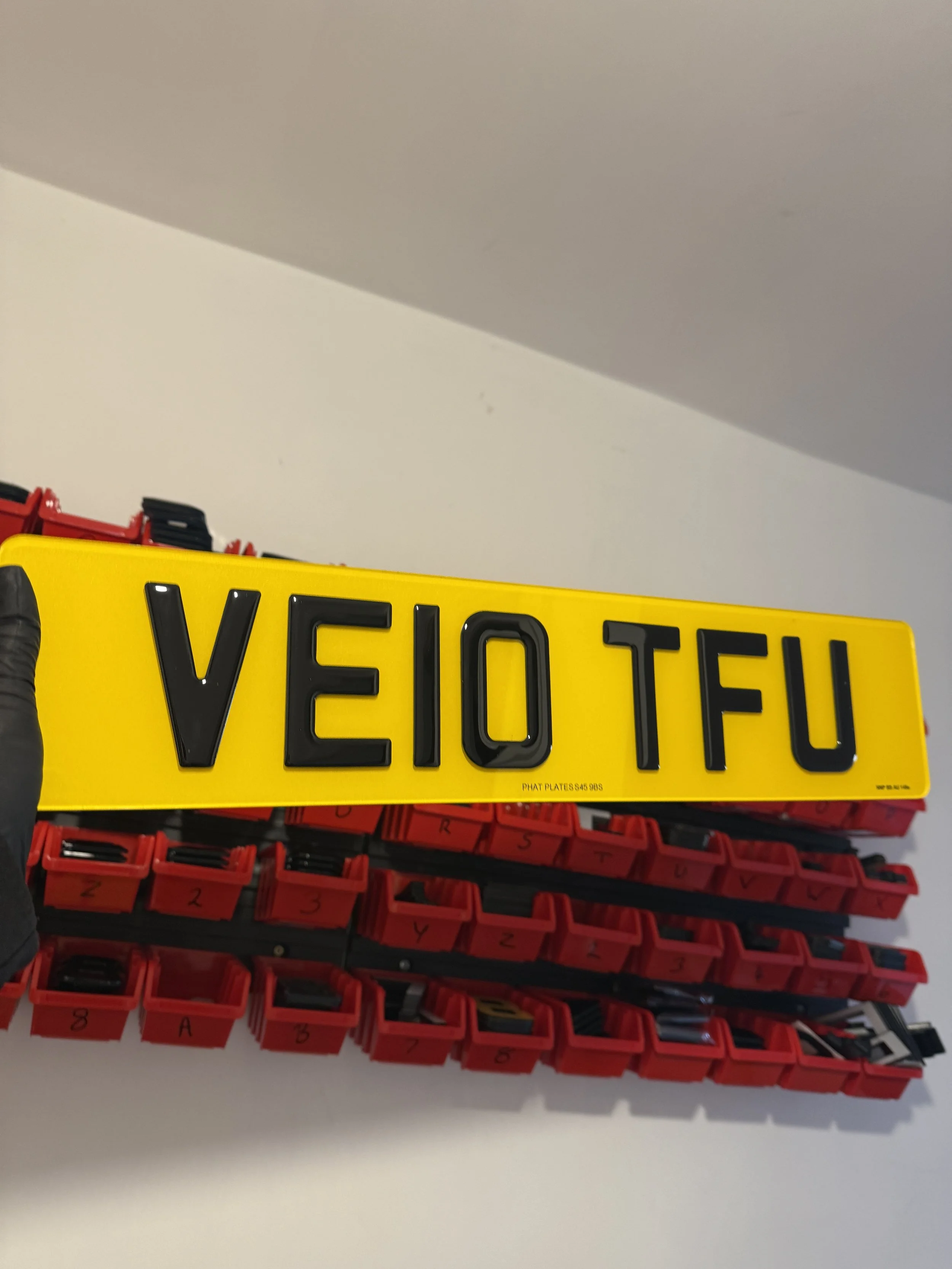 A person wearing a black glove holding a yellow license plate with black letters that spell 'VEIO TFU'. The background shows red storage bins stacked on top of each other.