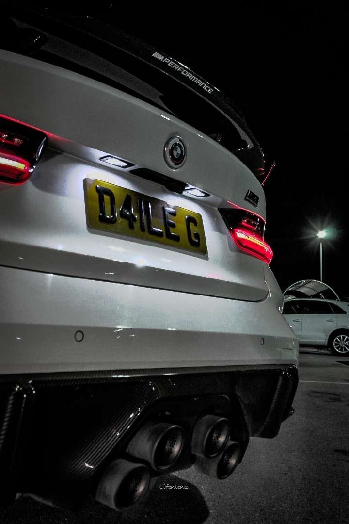 A white BMW car with a custom license plate reading 'D4LEC', parked in a dark parking lot at night, showing the rear view with taillights, diffuser, and quad exhaust pipes.