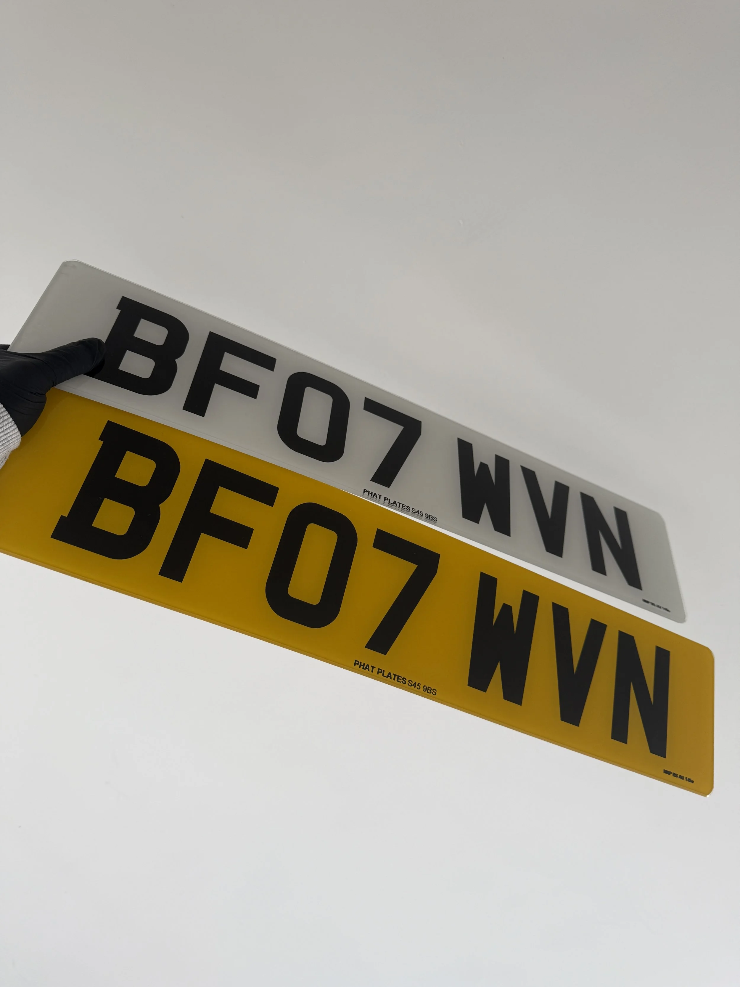 Two license plates, one white and one yellow, both with the same alphanumeric code 'BF07 WVN,' held against a gray sky, with a gloved hand visible holding the white plate.
