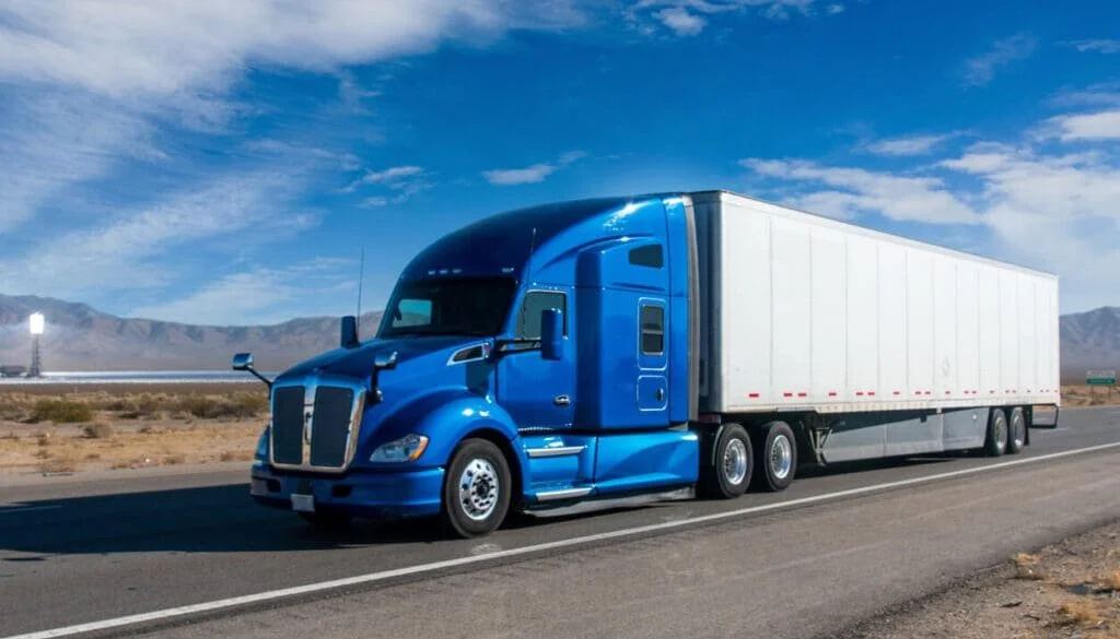 Blue semi-truck with white trailer driving on a highway through a desert landscape with mountains in the background.