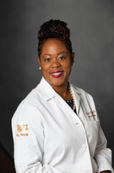 Portrait of an African American woman wearing a white medical coat with logo, smiling against a gray background.