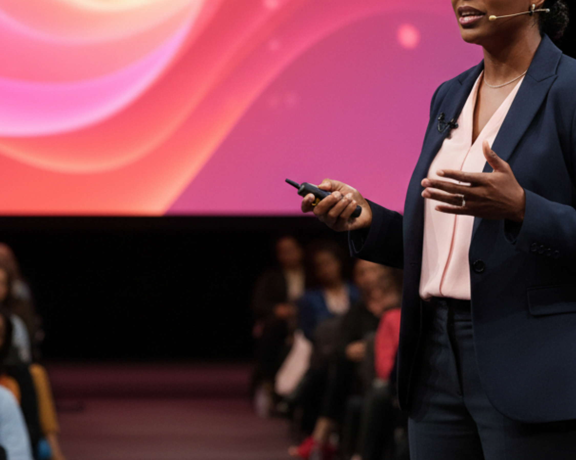 A woman giving a presentation at a conference or event, holding a remote control, wearing a navy blazer and pink blouse, with a large colorful screen behind her and an audience seated in front.