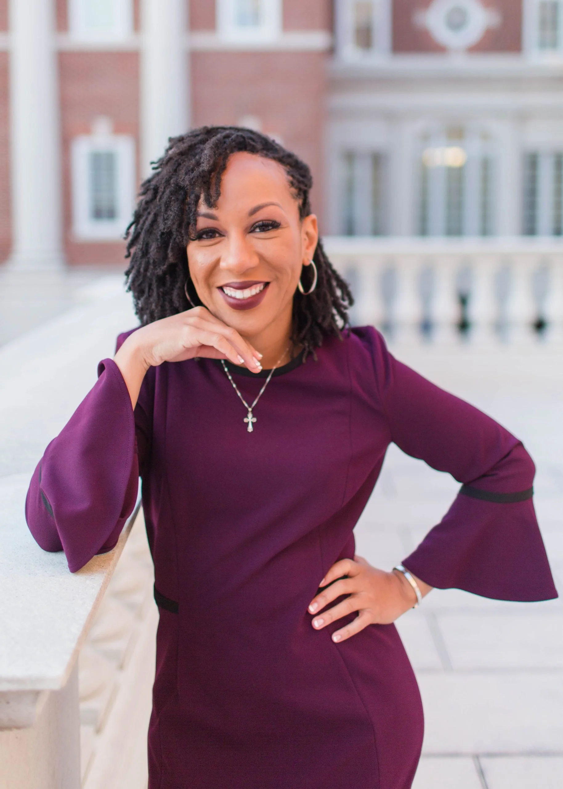 A smiling woman with dreadlocks, wearing a burgundy dress with bell sleeves, standing outdoors in front of a building with columns and a balcony.
