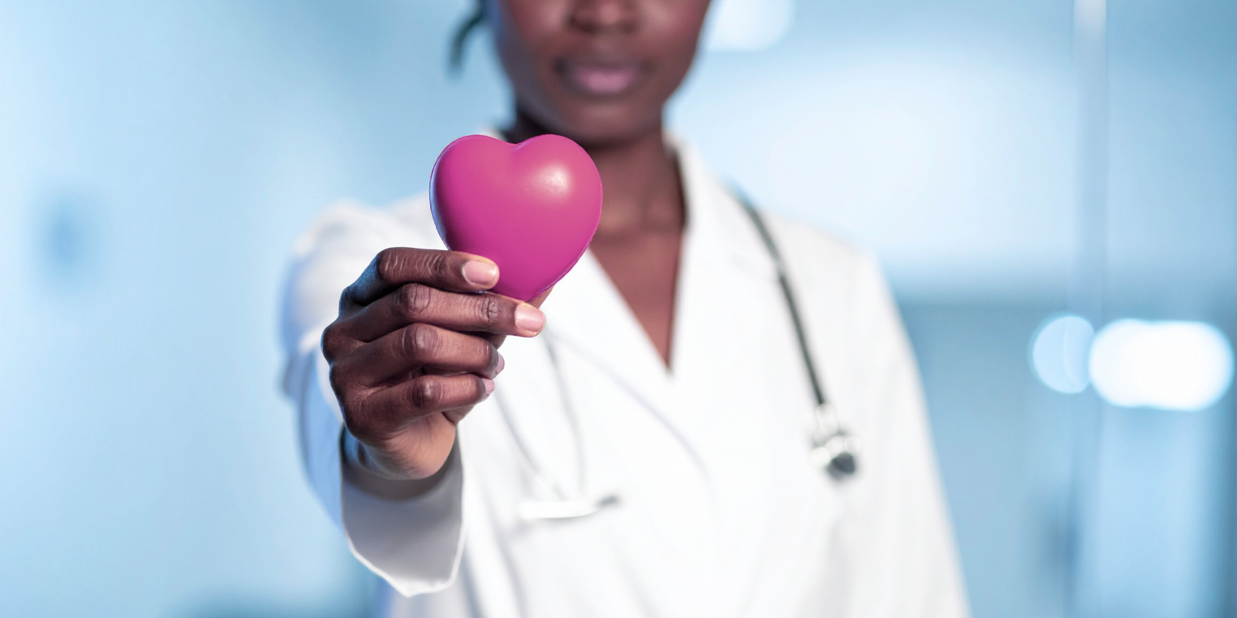 A healthcare professional holding a pink heart-shaped object, with a stethoscope around their neck, in a clinical setting.