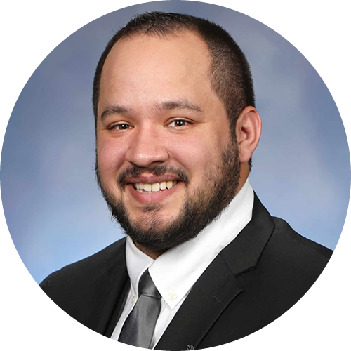 A man with short hair and a beard wearing a black suit, white shirt, and gray tie smiling against a blue background.