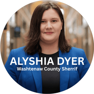 Portrait of Alyshia Dyer, Washtenaw County Sheriff, wearing a blue blazer and black shirt, standing outdoors with buildings in the background.