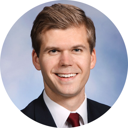 Portrait of a young man with light brown hair, smiling, wearing a black suit, white shirt, and red tie, with a light blue background.