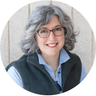 Portrait of a smiling woman with gray curly hair wearing glasses, a blue collared shirt, and a dark vest, sitting indoors with a light-colored wooden background.