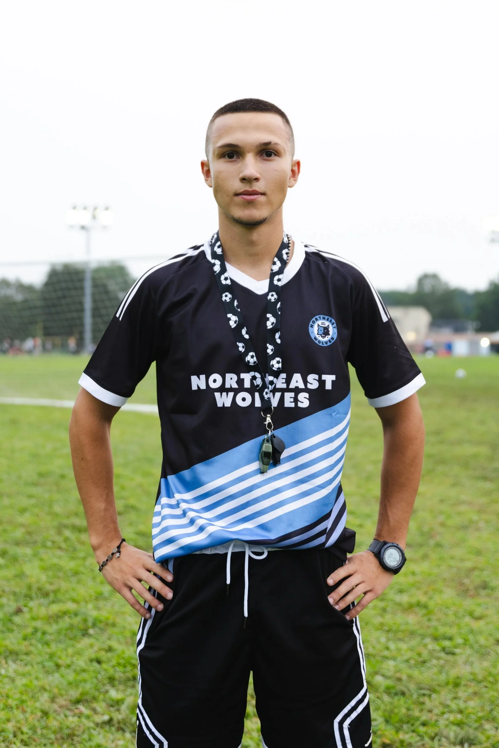A young male soccer player stands outdoors on a grassy field, wearing a black and blue soccer uniform with 'NORTHEAST WOLVES' written on the front. He has a whistle and a medal around his neck, a wristwatch, and has his hands on his hips.