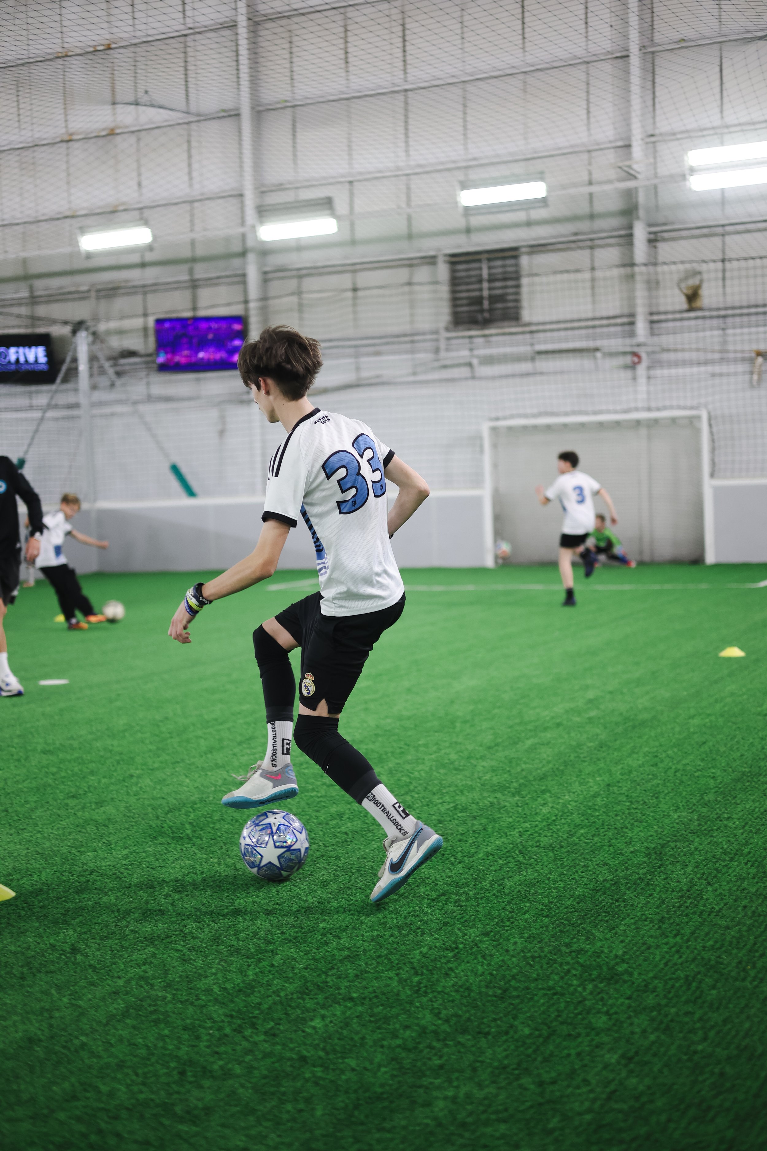 Young boy practicing soccer indoors on artificial turf, wearing a jersey with the number 33, dribbling a soccer ball, with other children and a coach in the background.