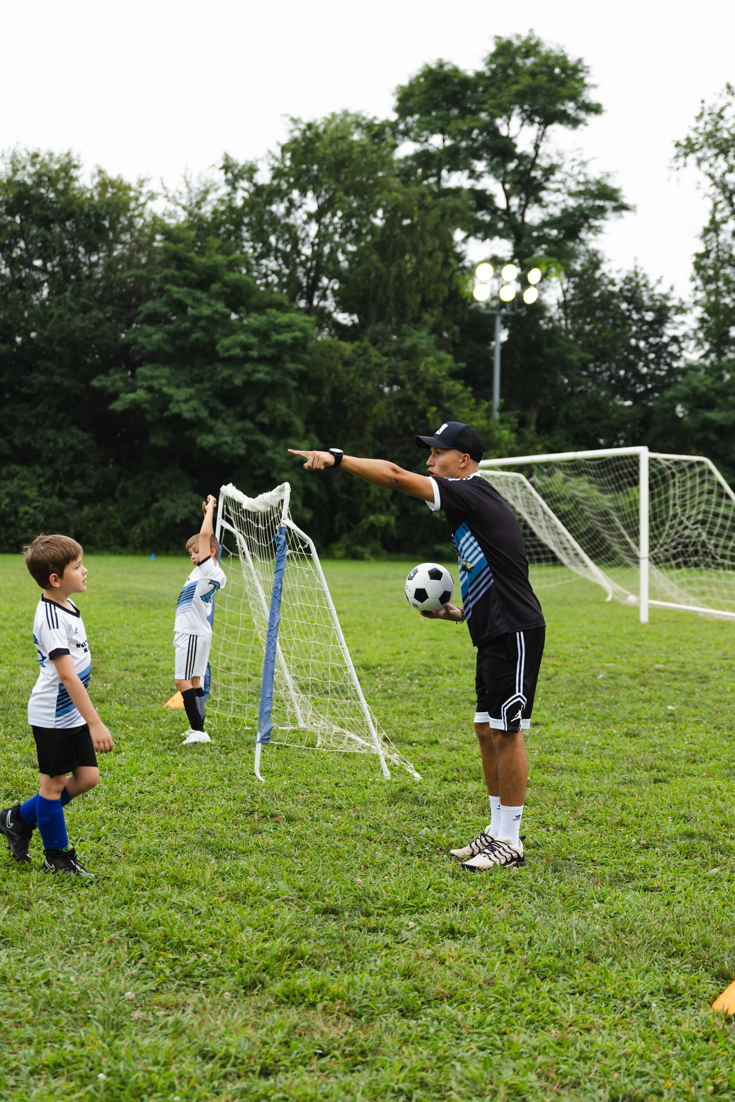 A coach giving instructions to young children during a soccer practice on a grassy field with mini goals and trees in the background.