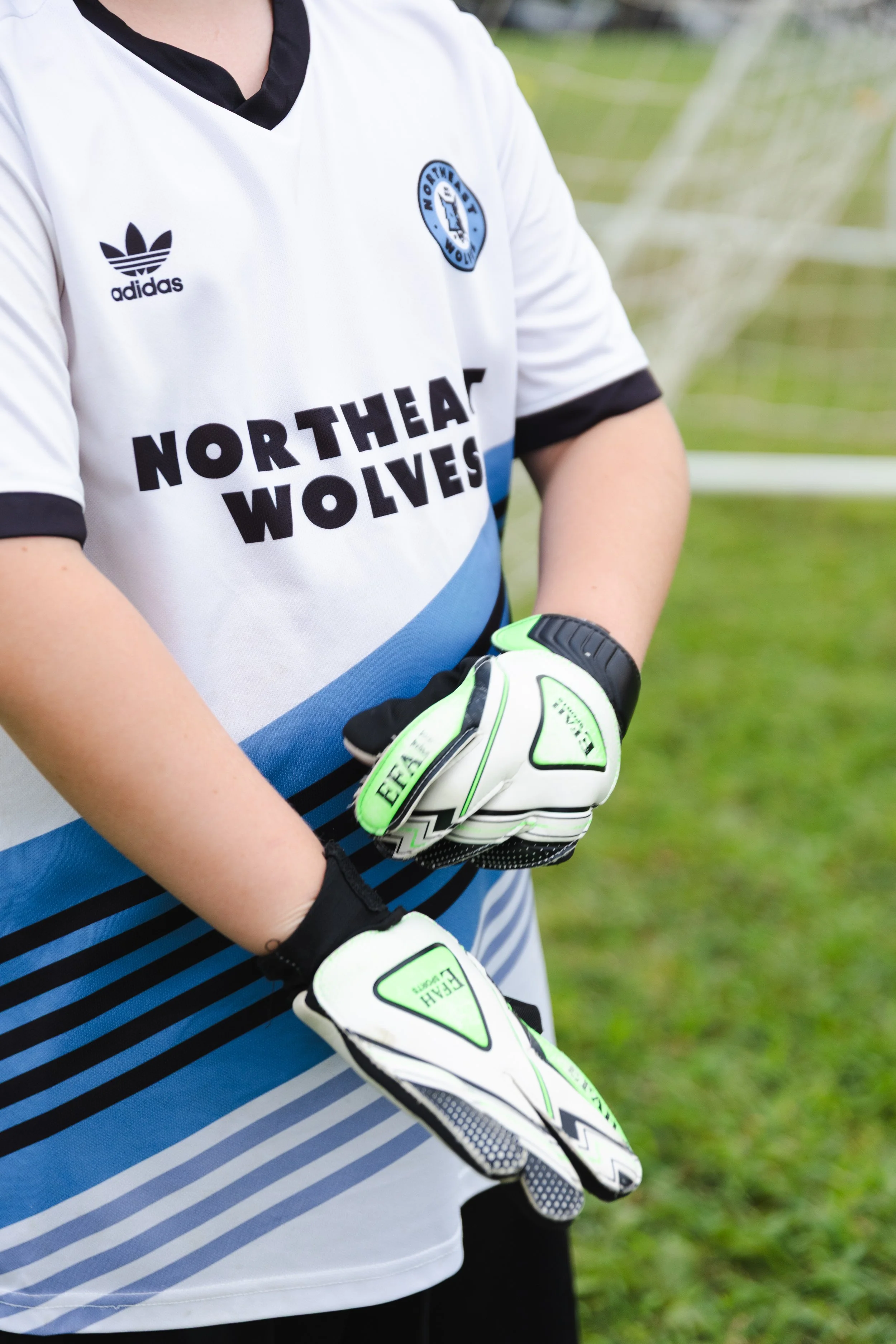 A soccer player wearing a white jersey with black and blue accents, labeled 'Northeast Wolves,' and black and neon green goalkeeper gloves, standing on a grass field with a goal net in the background.