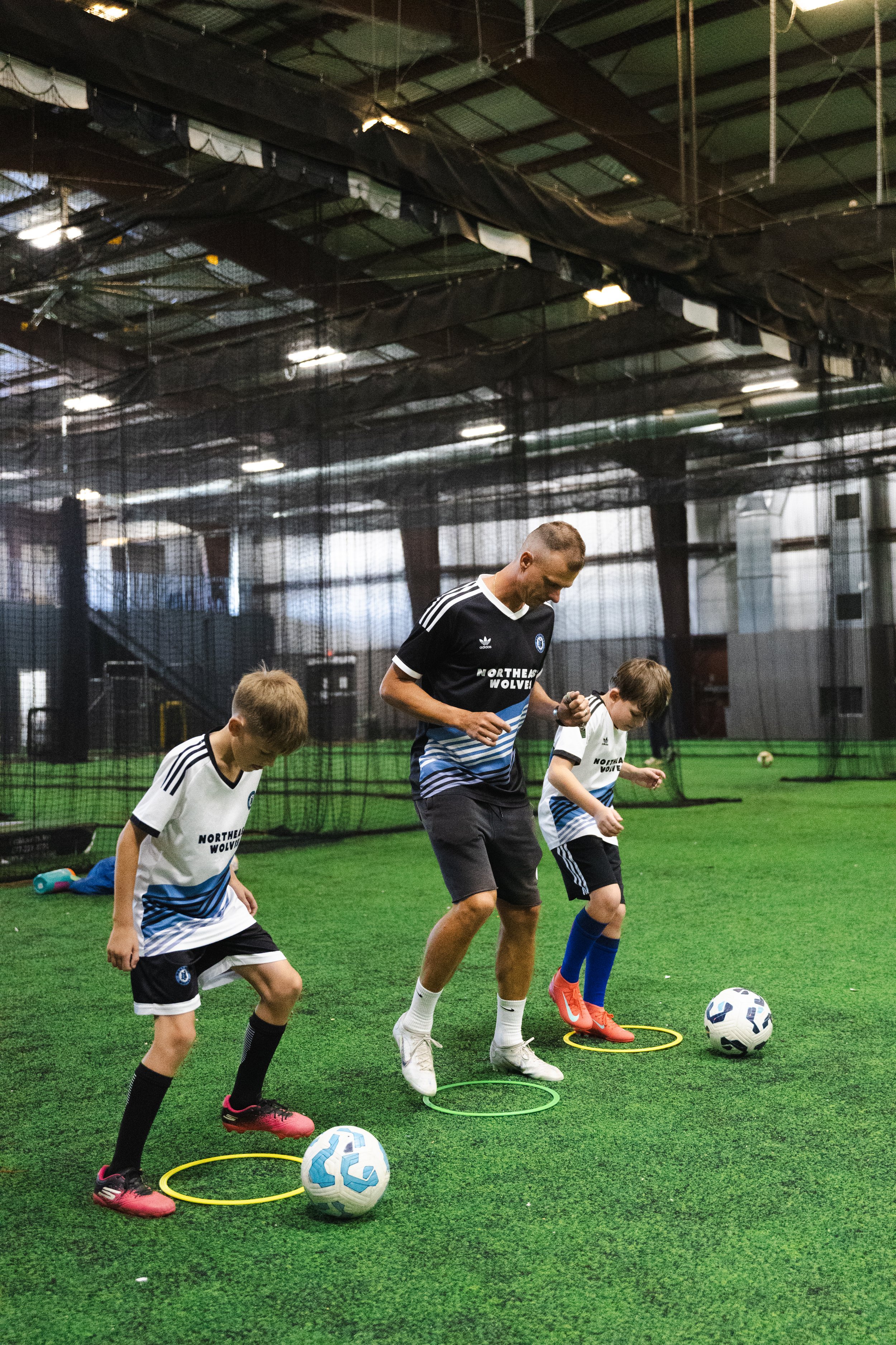 A coach training two young boys in soccer drills inside an indoor soccer facility, with each boy passing through a circle and practicing with a soccer ball.