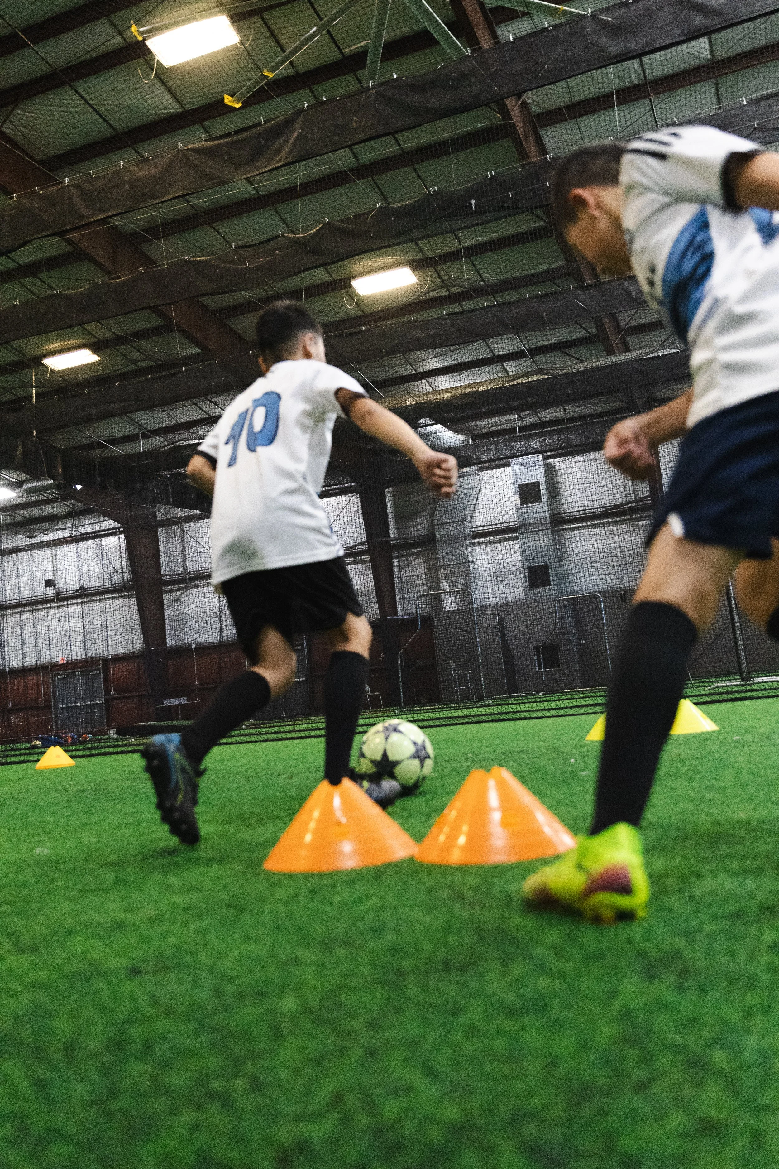 Two young soccer players during an indoor training session, dribbling a ball between orange cones on artificial turf.