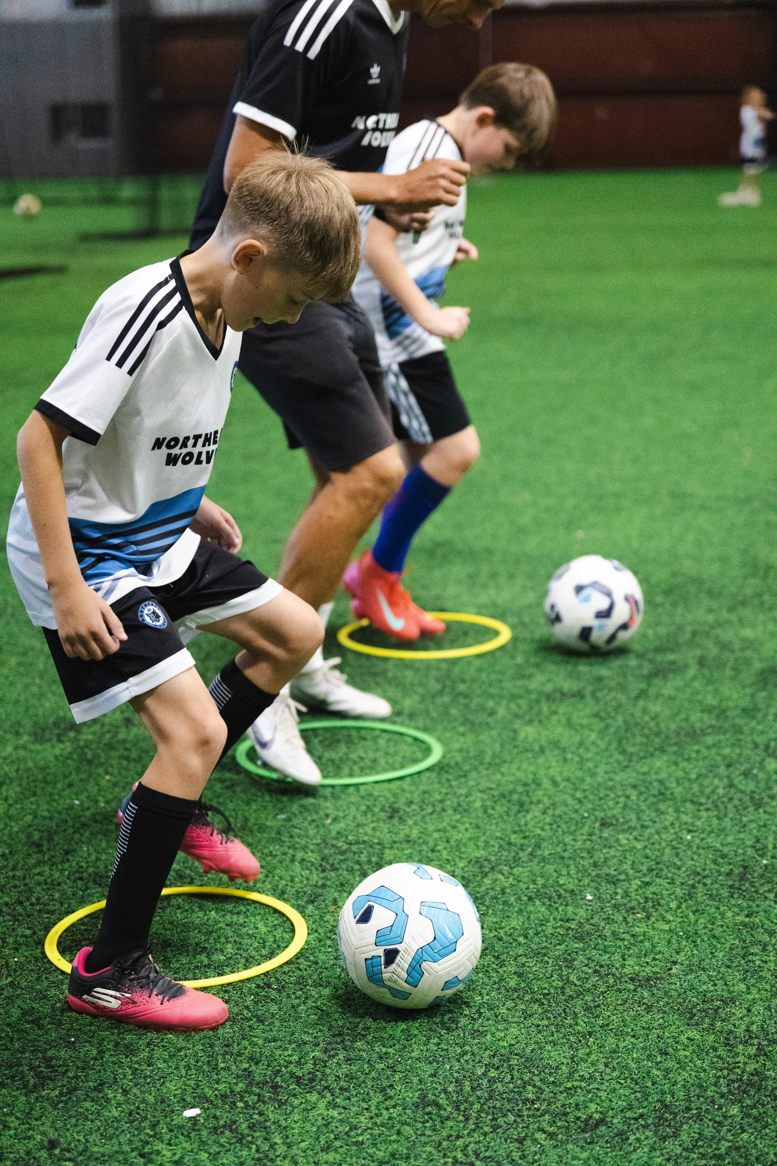 Young boys practicing soccer drills indoors, stepping through colorful hoops on artificial turf, with soccer balls nearby, supervised by an adult coach.