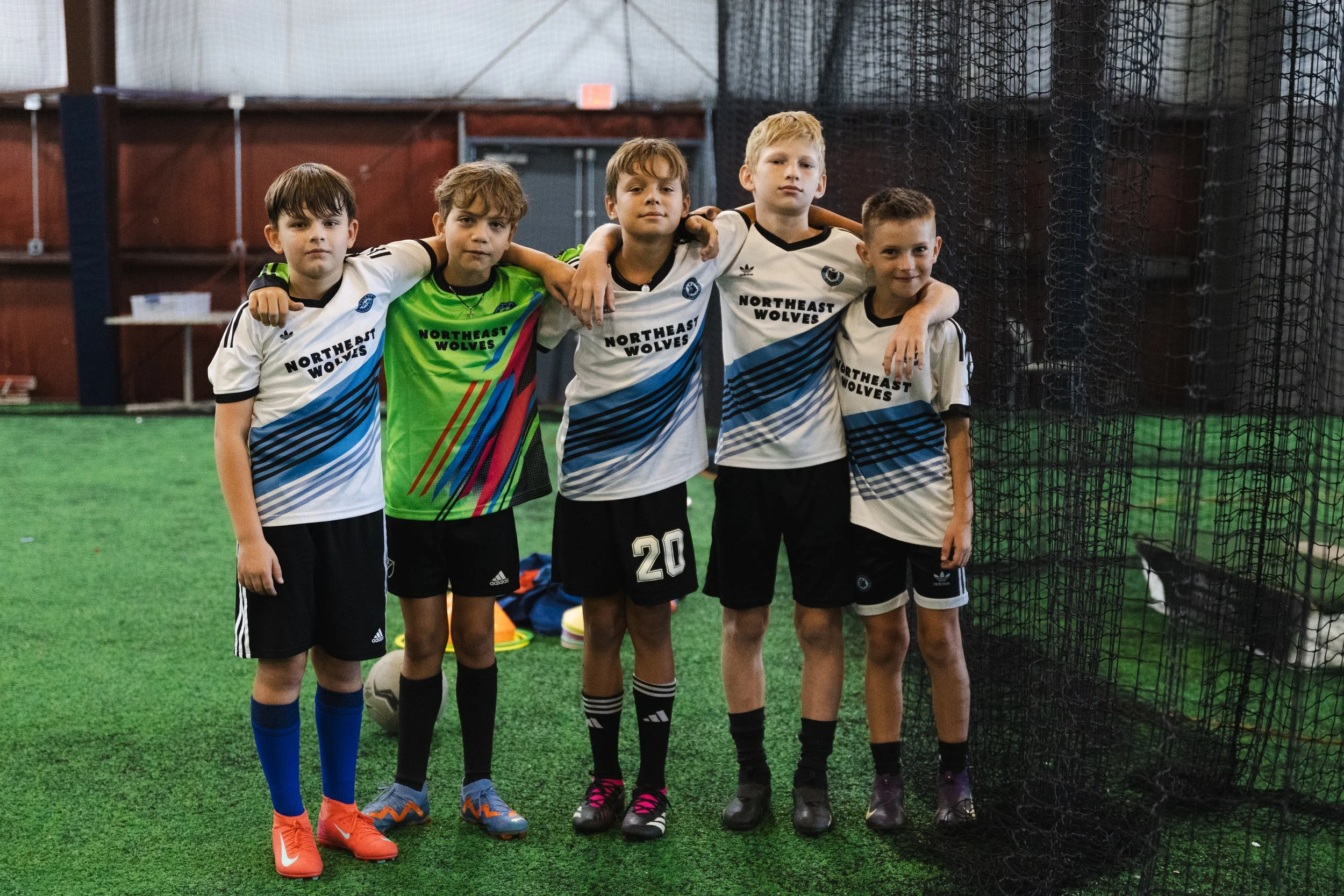 Five young boys posing together indoors on a football field, dressed in sports uniforms, with arms around each other, standing near a black net.