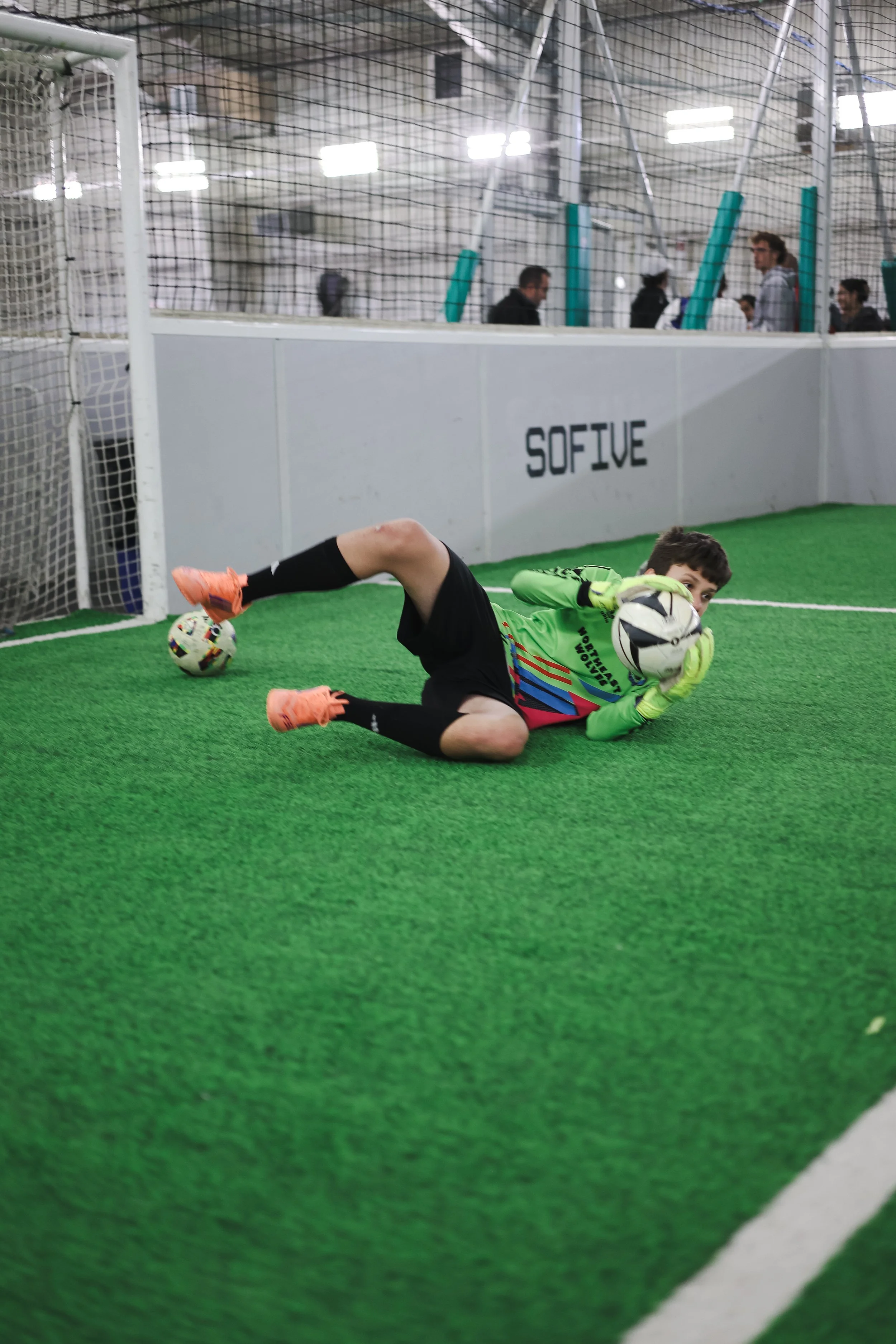 A young boy wearing a green goalkeeper jersey and orange shoes making a save during indoor soccer, lying on artificial green turf with a soccer ball near his hand.
